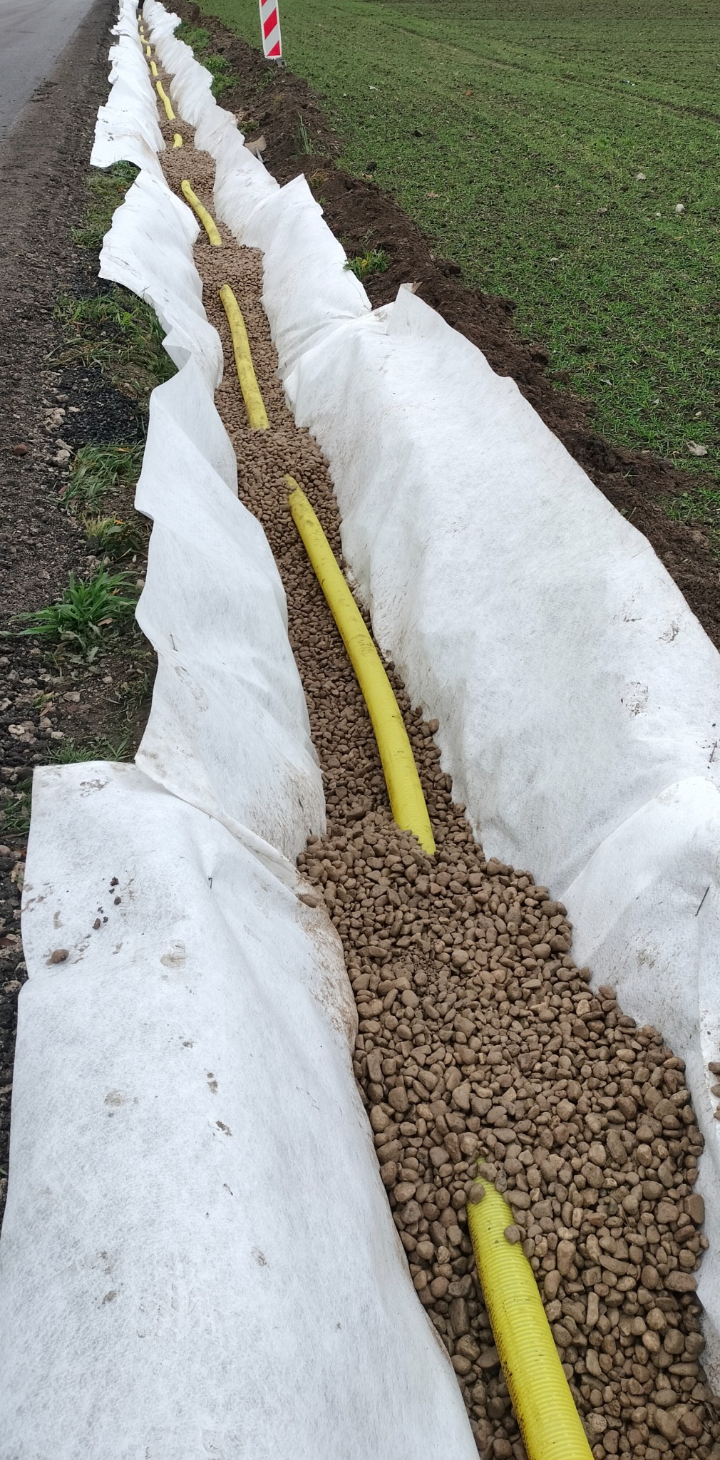 Drenaż liniowy z żółtą rurą drenażową, obsypany żwirem i wyłożony białą geowłókniną, widok z góry wzdłuż wykopu.