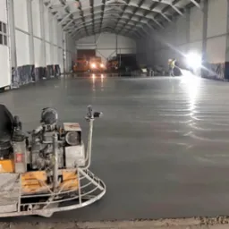 Zacieraczka mechaniczna do betonu w trakcie pracy na dużej powierzchni hali przemysłowej, w tle pracownik spryskujący podłoże i oświetlenie robocze.