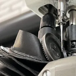 Zbliżenie na igłę maszyny do szycia przeszywającej warstwy grubego, czarnego materiału, prawdopodobnie skórzanego, z widocznymi nitkami i mechanizmem dociskowym.