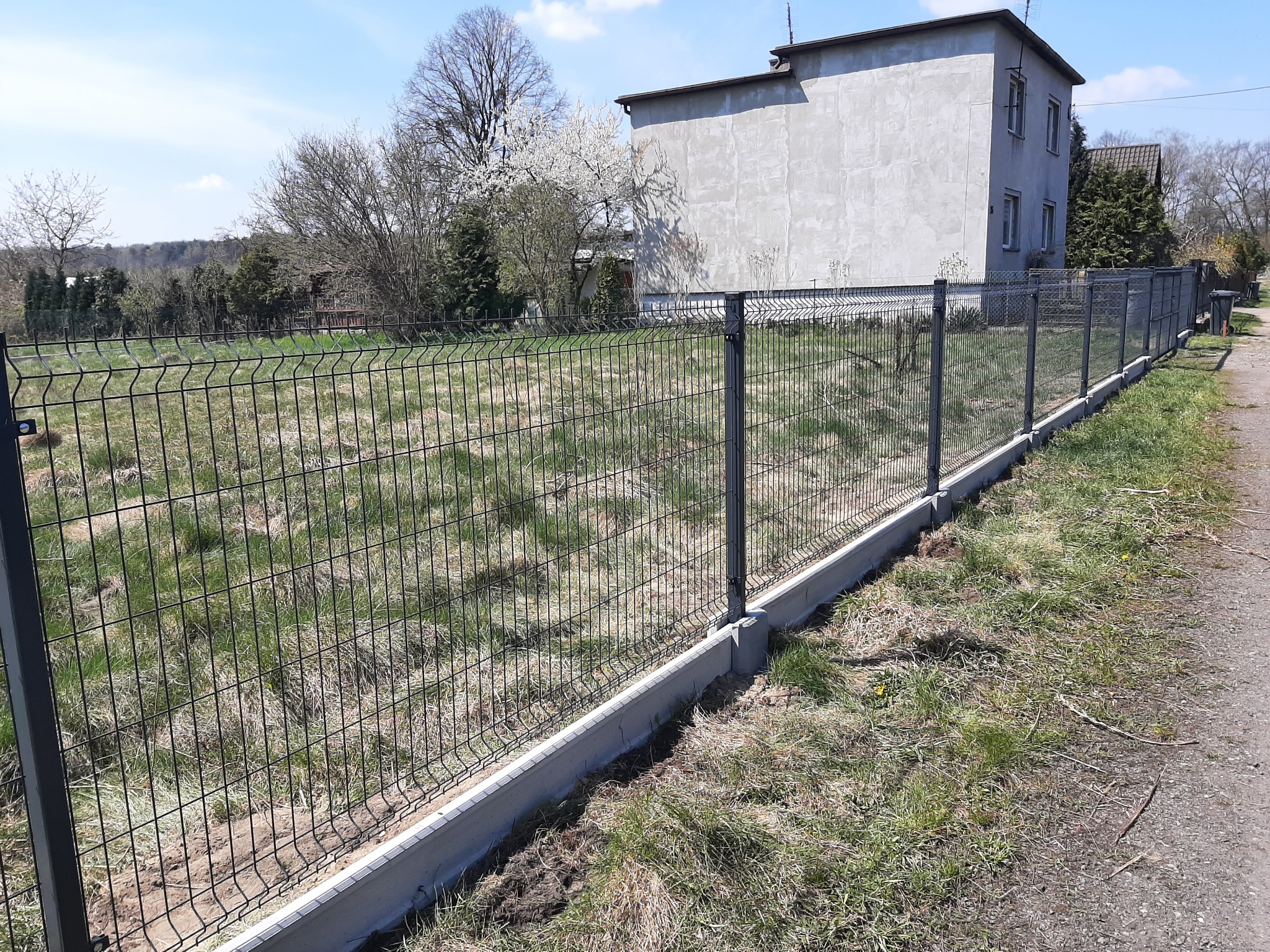 Długi, czarny panel ogrodzeniowy z betonową podmurówką, widok z zewnątrz posesji, w tle budynek mieszkalny i drzewa.