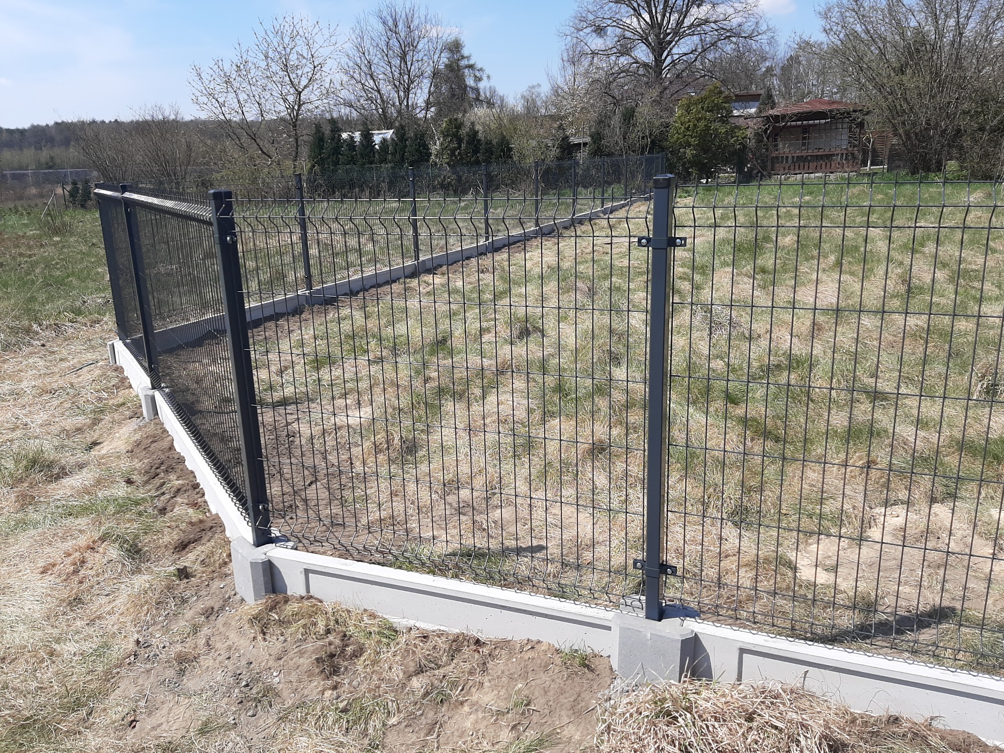 Nowoczesne ogrodzenie panelowe z podmurówką betonową, widok na łąkę i zabudowania w tle.
