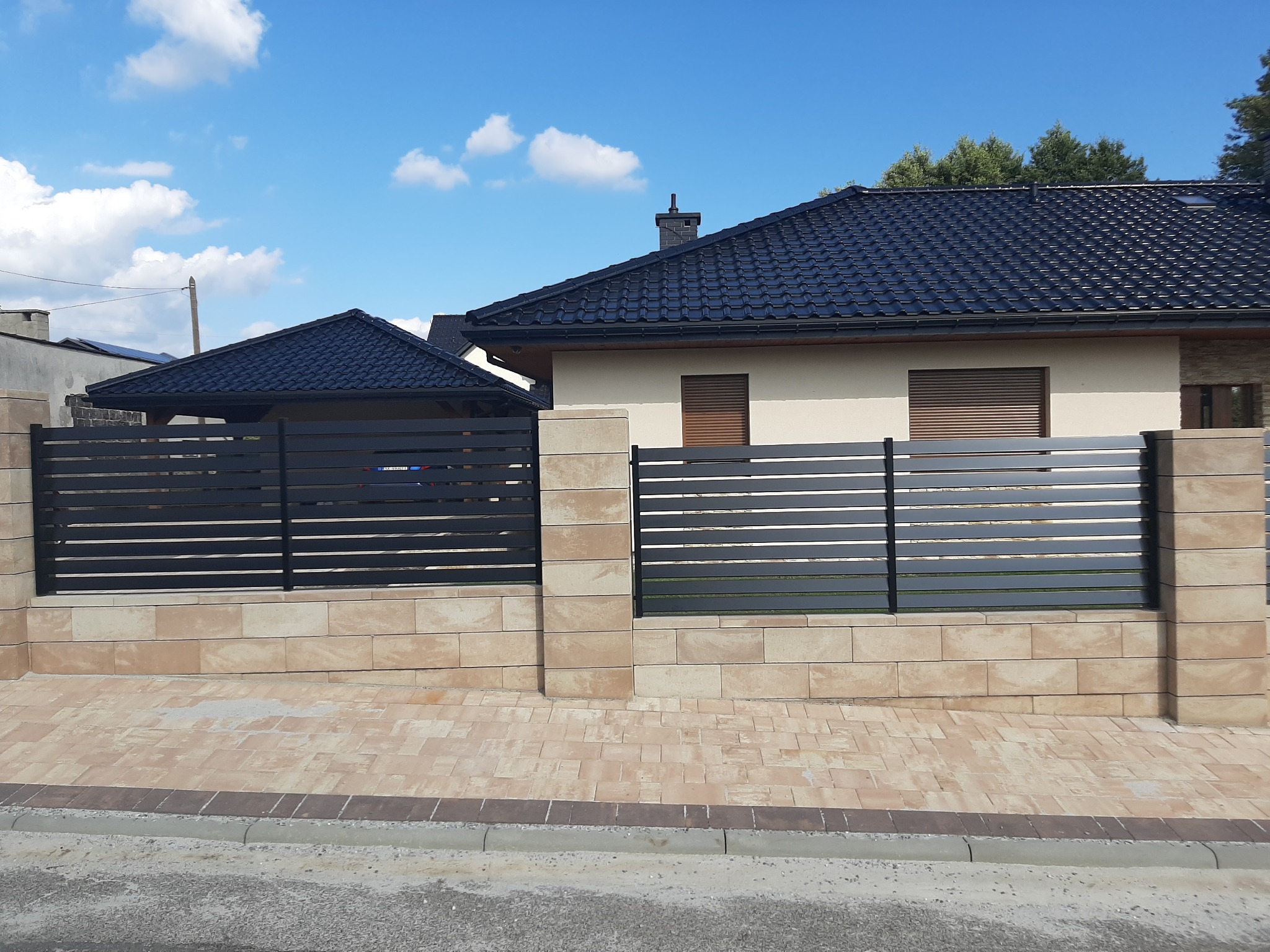 Nowoczesne ogrodzenie z poziomych, grafitowych paneli zamontowane na niskim murku z beżowych bloczków, na tle domu z ciemną dachówką i błękitnego nieba.