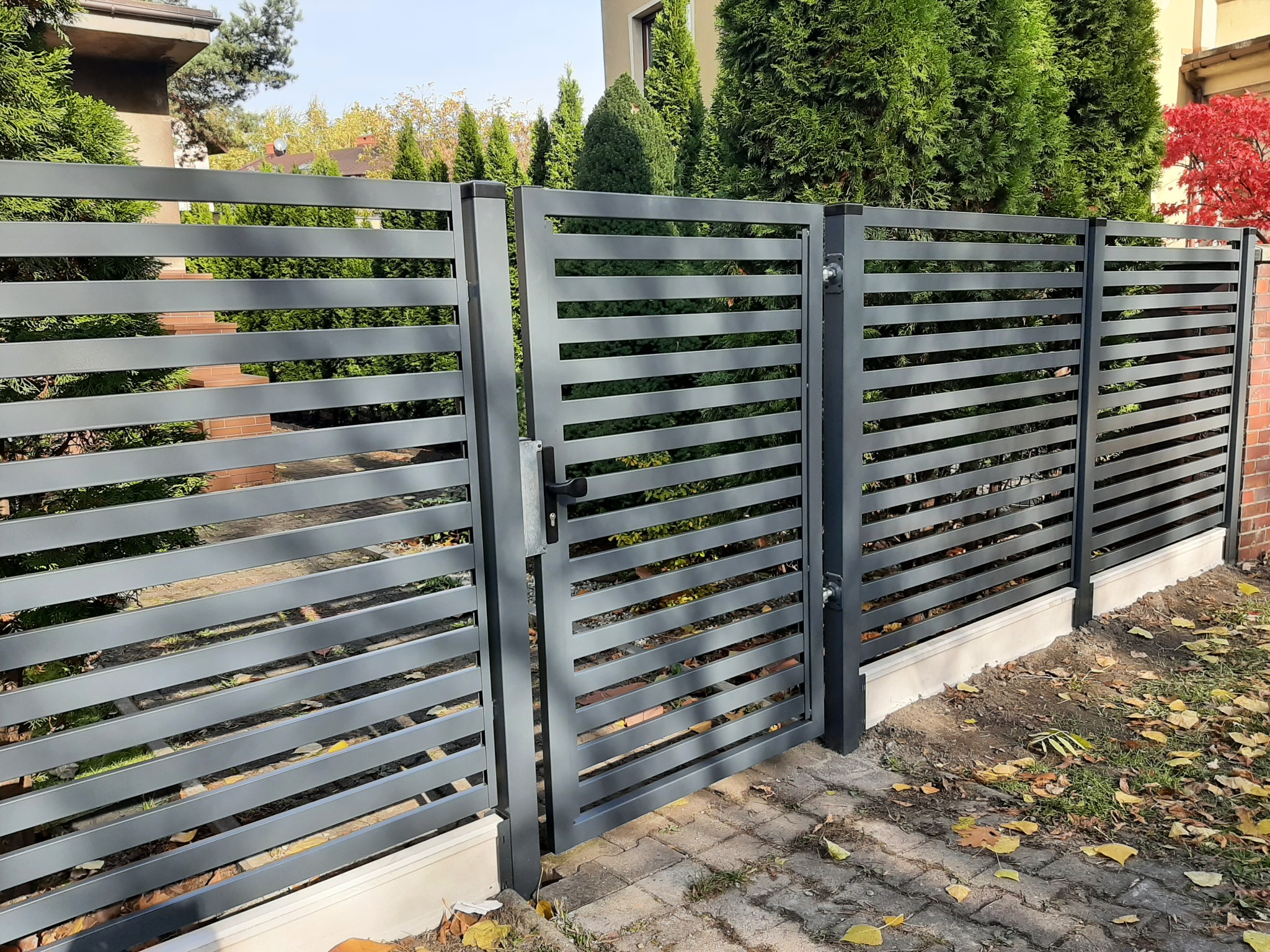 Nowoczesne, poziome przęsła ogrodzenia i furtka w kolorze antracytowym, zamontowane w Rudzie Śląskiej, z widocznymi nasadzeniami i brukowaną ścieżką.