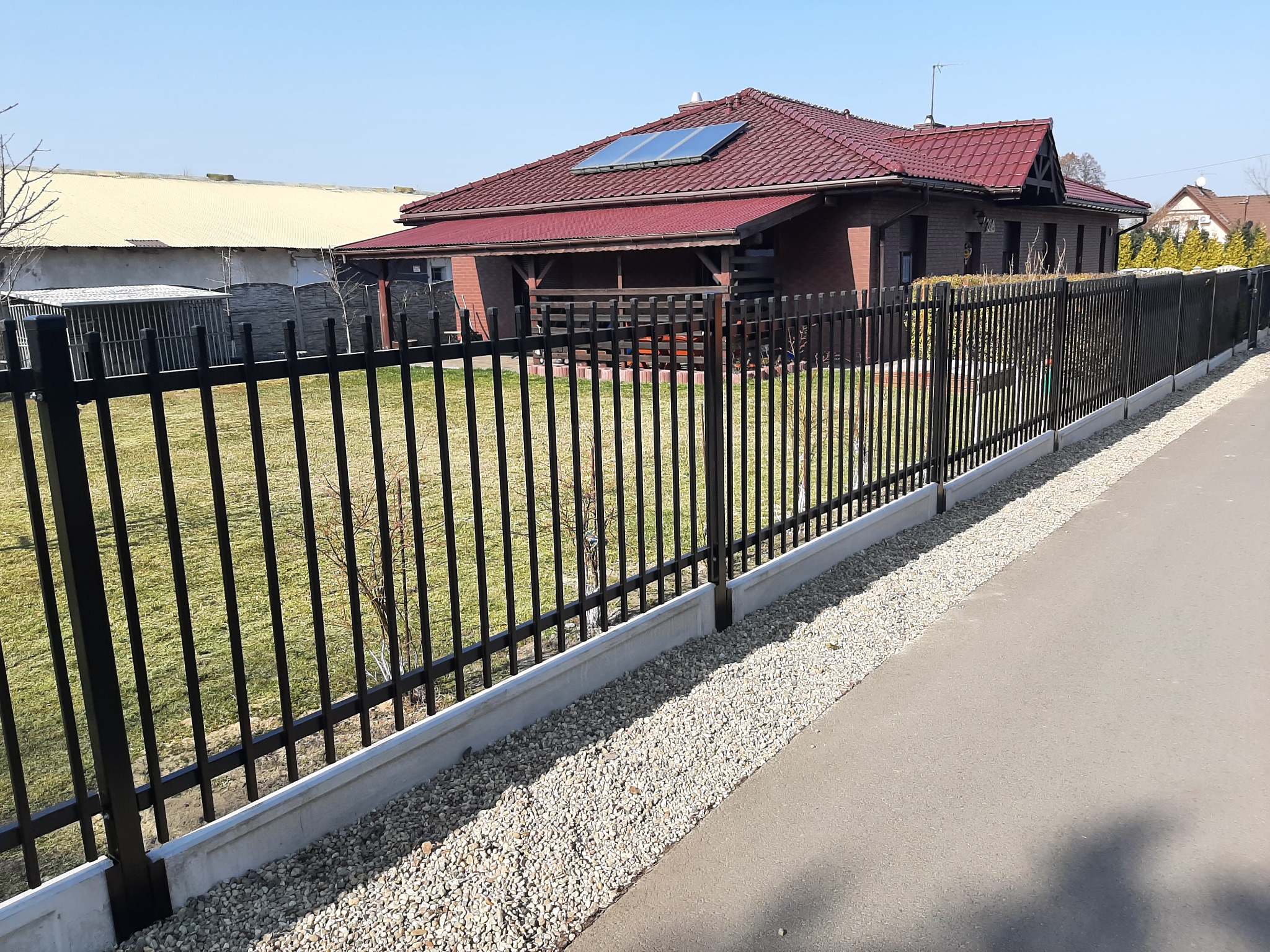 Nowoczesne ogrodzenie metalowe z pionowych prętów, osadzone na betonowej podmurówce, z dekoracyjnym obsypaniem z jasnych kamyków, na tle domu z czerwoną dachówką i panelem solarnym.