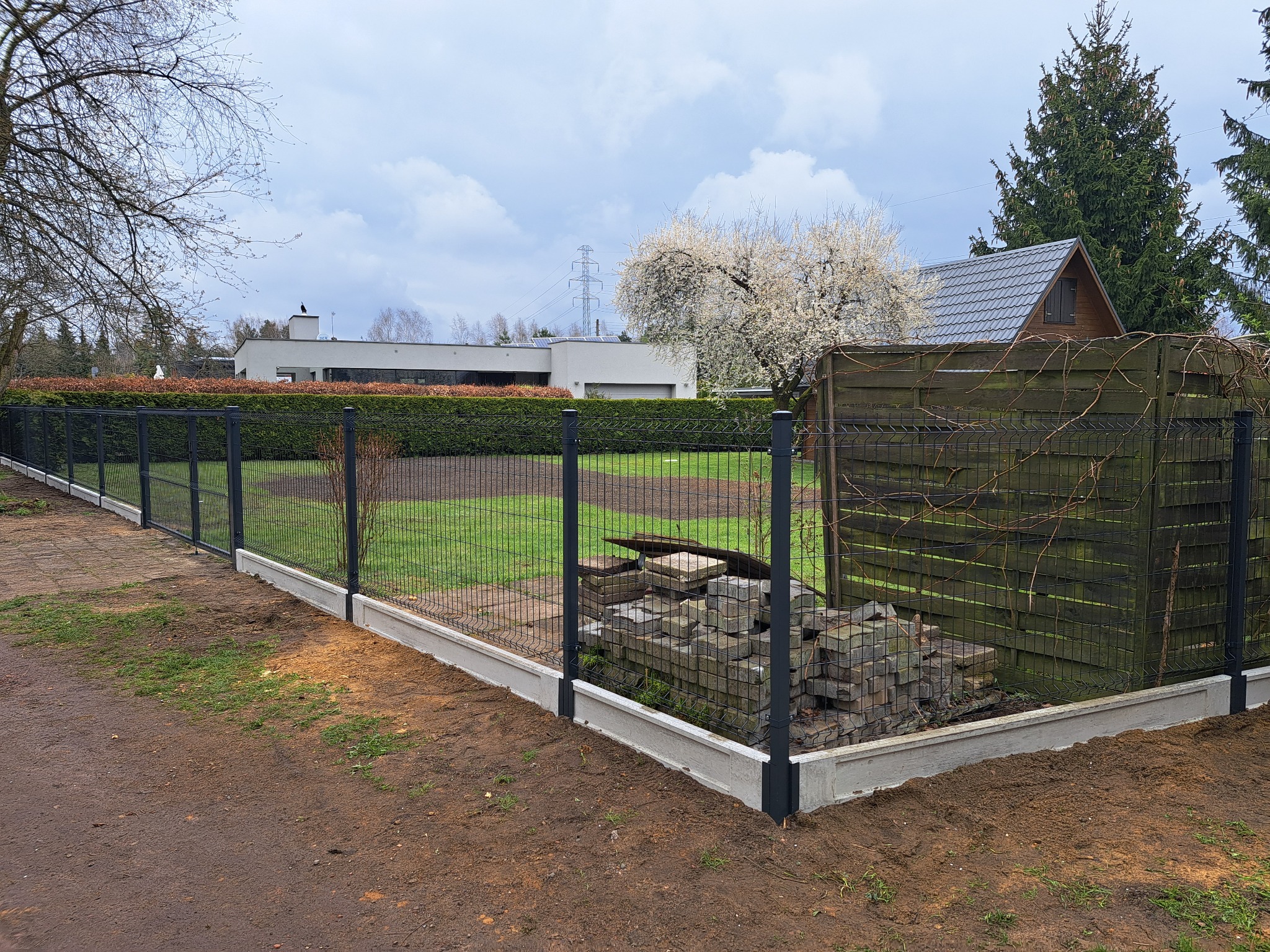 Czarny panelowy płot z betonową podmurówką na nierównym, ziemistym terenie, z widocznym stosem kostki brukowej za ogrodzeniem i domami w tle.