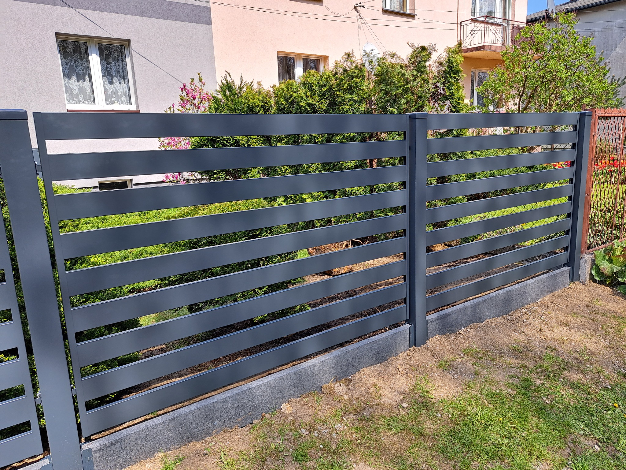 Nowoczesne, poziome ogrodzenie metalowe w kolorze grafitowym, z betonową podmurówką, na tle zieleni ogrodu i elewacji domu.