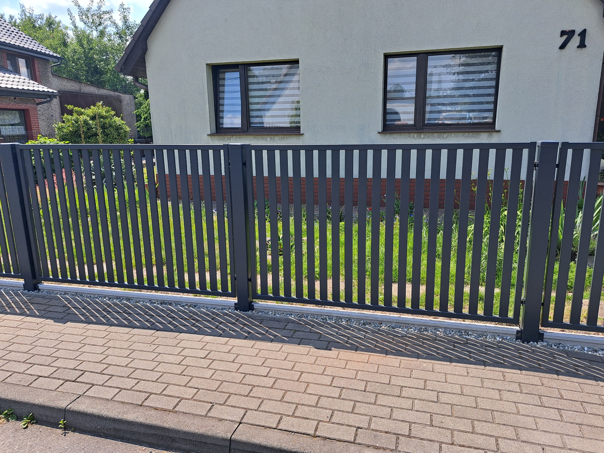 Nowoczesne, grafitowe ogrodzenie z pionowych paneli na podmurówce z drobnych kamieni, z widokiem na dom z numerem 71 i oknami z żaluzjami poziomymi.