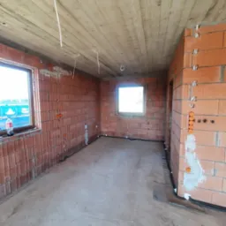 Surowy stan wnętrza budynku w trakcie prac elektrycznych: widoczne przewody wystające z sufitu, puszki instalacyjne w ścianach z cegły dziurawki, okno z butelką Coca-Coli na parapecie i fragment...