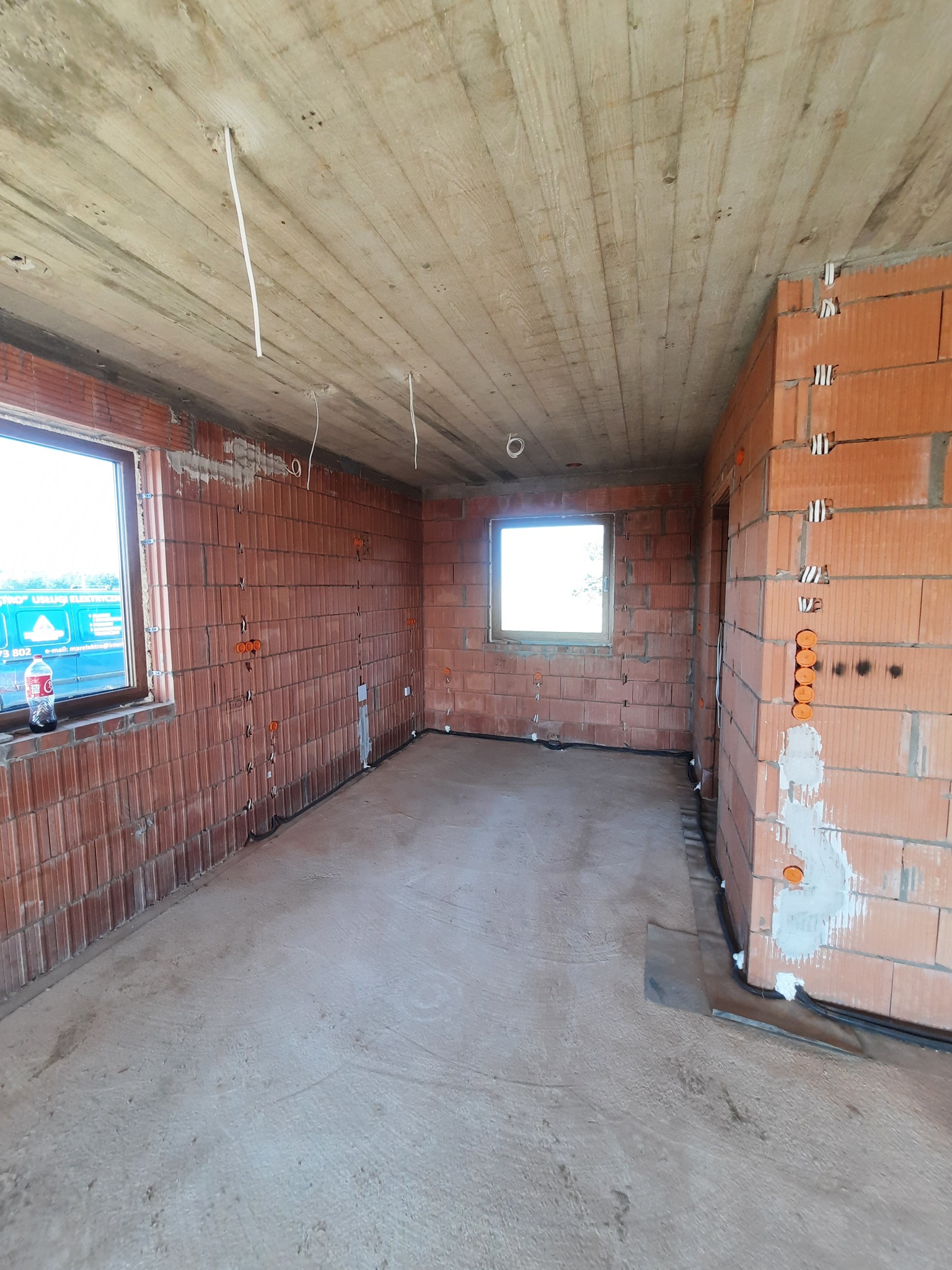 Surowy stan wnętrza budynku w trakcie prac elektrycznych: widoczne przewody wystające z sufitu, puszki instalacyjne w ścianach z cegły dziurawki, okno z butelką Coca-Coli na parapecie i fragment...