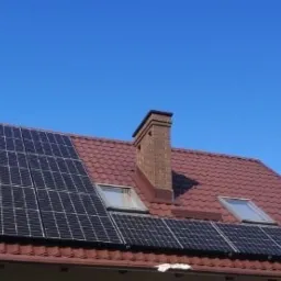 Panele fotowoltaiczne na dachu domu z czerwoną dachówką i ceglanym kominem, widoczne okna dachowe.