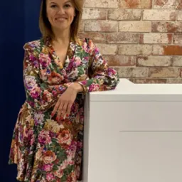 Uśmiechnięta kobieta w kwiecistej sukience, oparta o białe biurko na tle ceglanej ściany w biurze kancelarii prawnej.