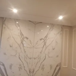 Ściana wyłożona dużymi płytami z białego marmuru z szarymi żyłami, symetryczny wzór, oświetlenie punktowe, dekoracyjne listwy ścienne.