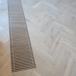 Jasne, parkietowe panele ułożone we wzór jodełki francuskiej z długą, wąską kratką wentylacyjną w podłodze.