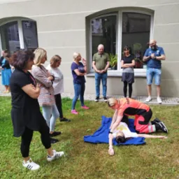 Instruktor w stroju ratowniczym demonstruje resuscytację krążeniowo-oddechową na manekinie na trawie, grupa osób obserwuje z uwagą, budynek w tle.