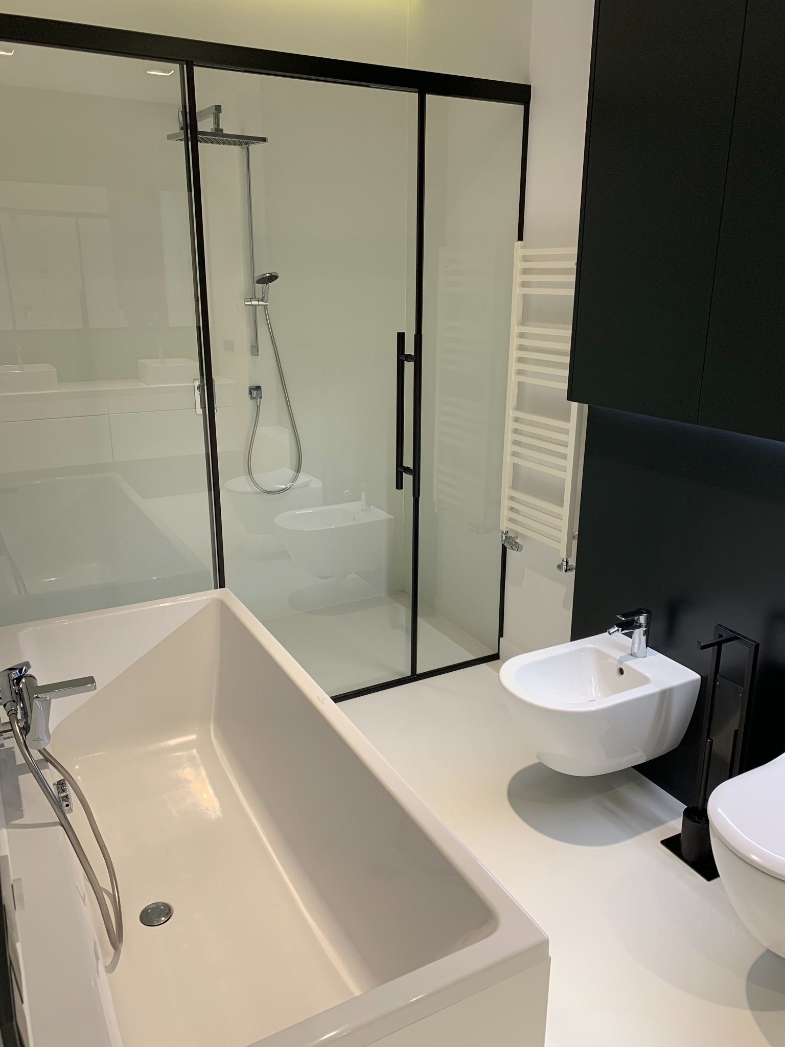 Nowoczesna łazienka w bieli i czerni z wanną, bidetem i kabiną prysznicową z czarnymi ramami, jasna podłoga i biały grzejnik drabinkowy.