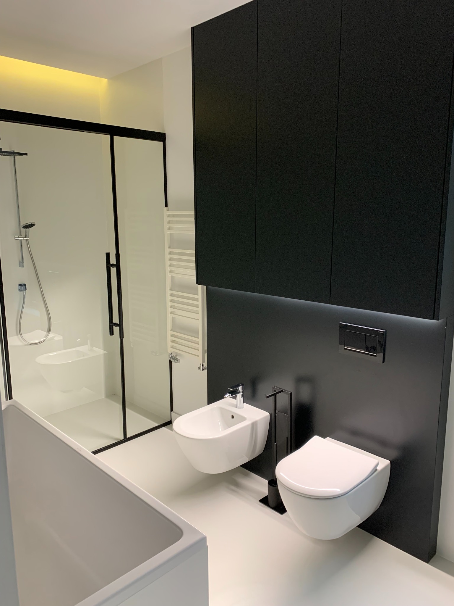 Minimalistyczna łazienka w bieli i czerni: kabina prysznicowa z czarną ramą, podwieszana toaleta i bidet, czarne szafki wiszące z podświetleniem LED, biały grzejnik drabinkowy.