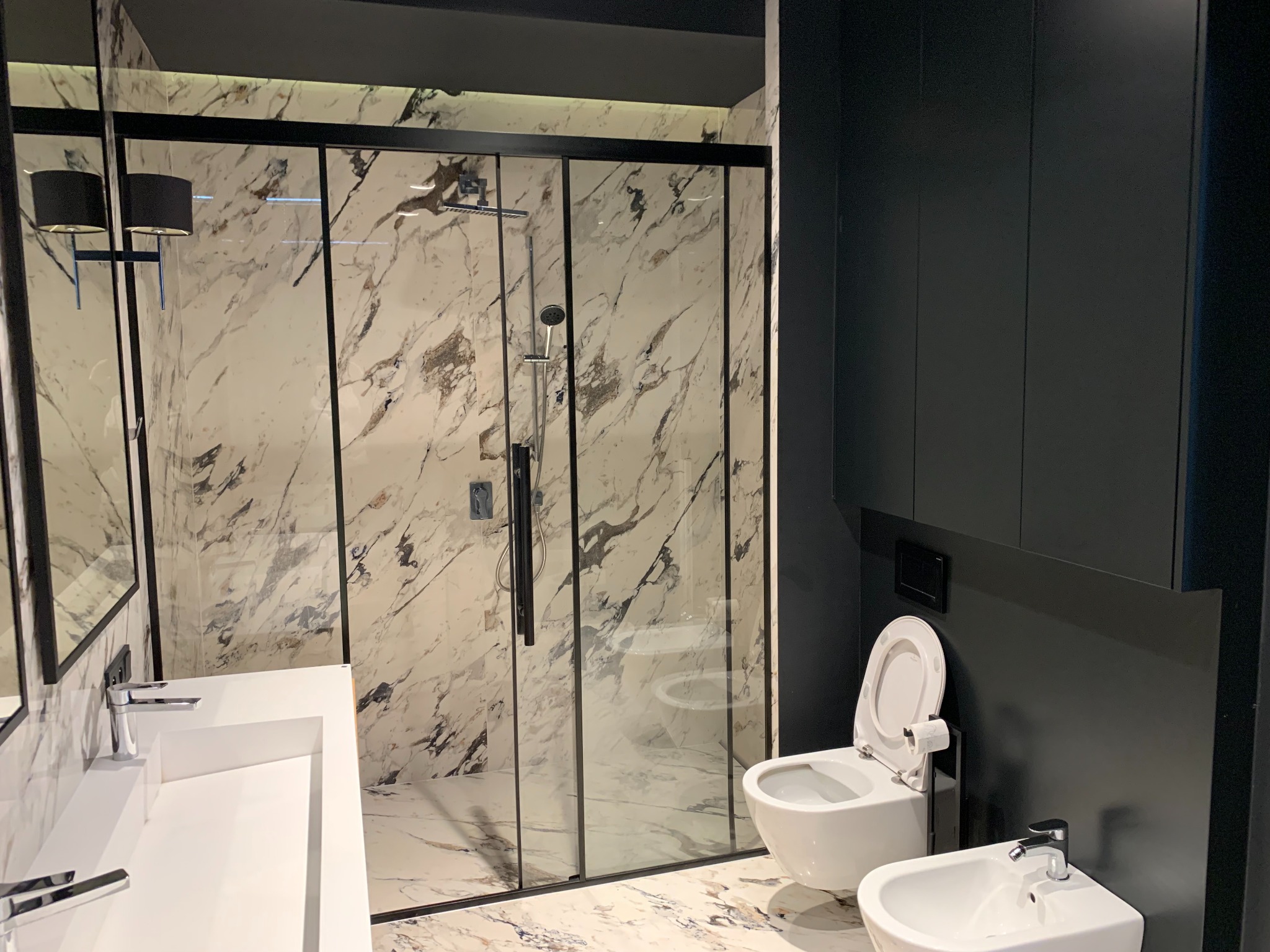 Nowoczesna łazienka z kabiną prysznicową typu walk-in, umywalką nablatową i wiszącą toaletą oraz bidetem, wykończona płytkami imitującymi marmur i czarnymi elementami.