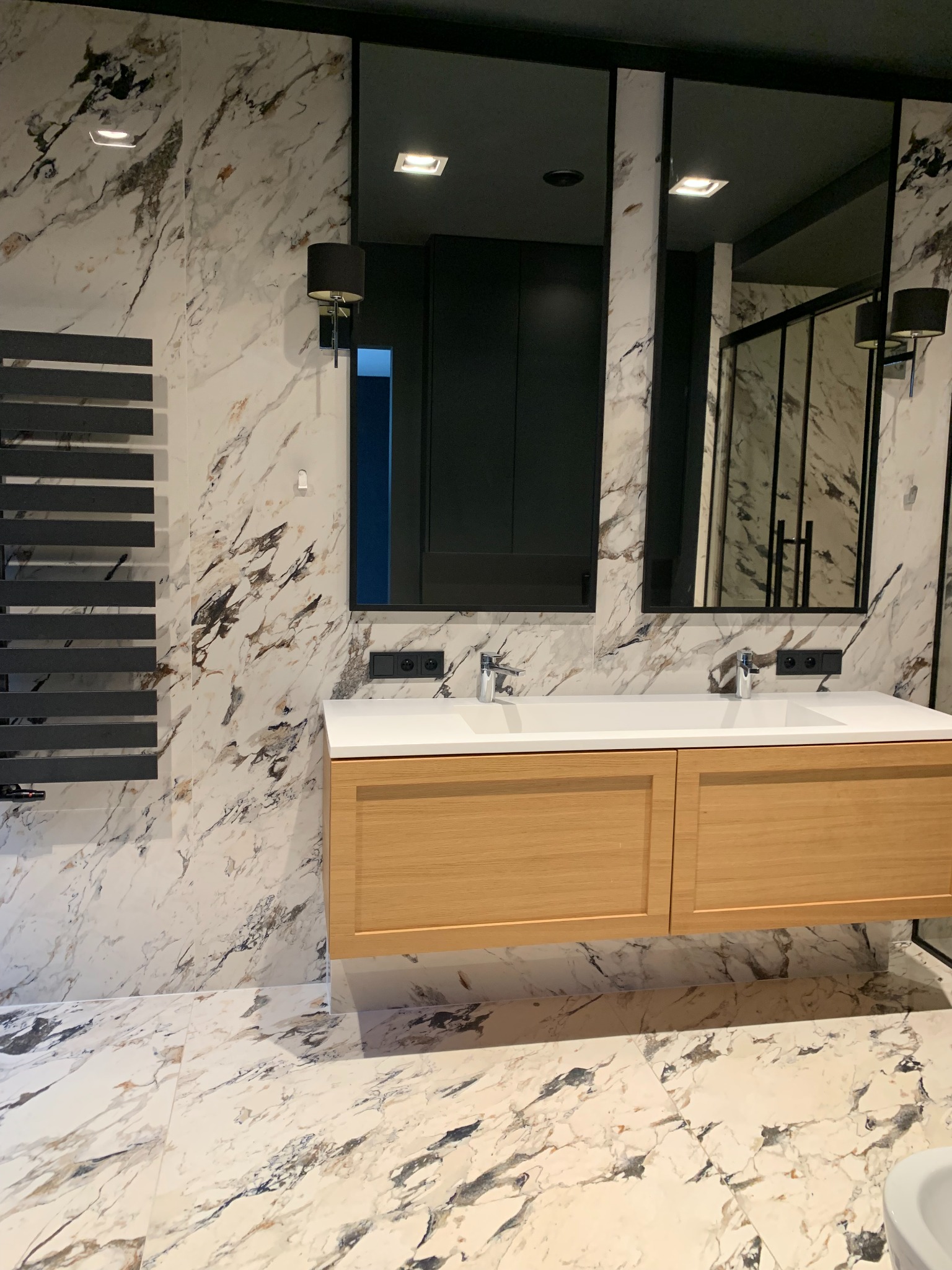 Nowoczesna łazienka z marmurowymi płytkami na ścianach i podłodze, drewniana szafka pod umywalką z dwoma chromowanymi kranami, czarne lustra i grzejnik drabinkowy.