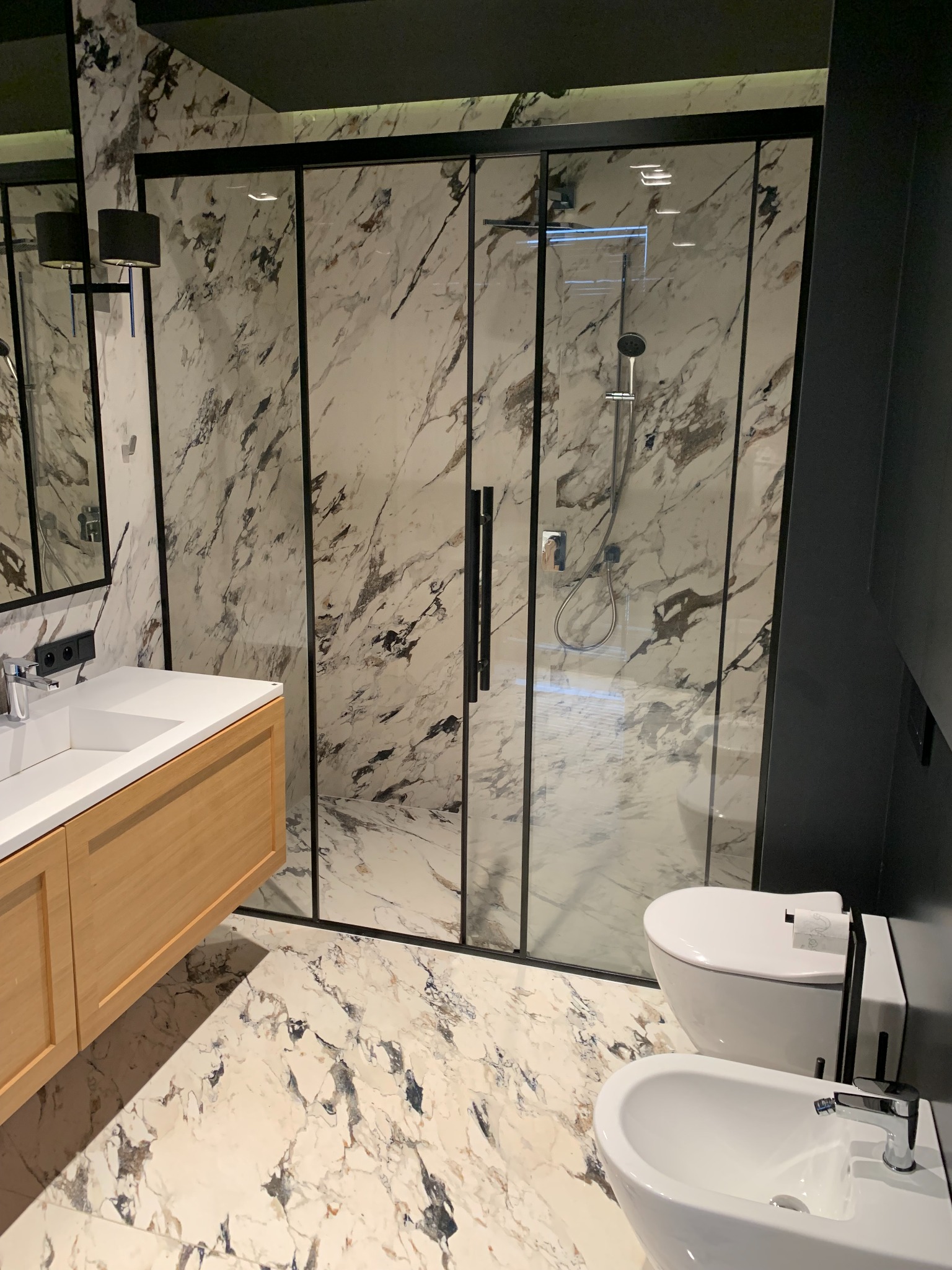 Nowoczesna łazienka z kabiną prysznicową z czarnymi ramami, marmurowymi płytkami na ścianach i podłodze, drewnianą szafką pod umywalką oraz wiszącą toaletą i bidetem.