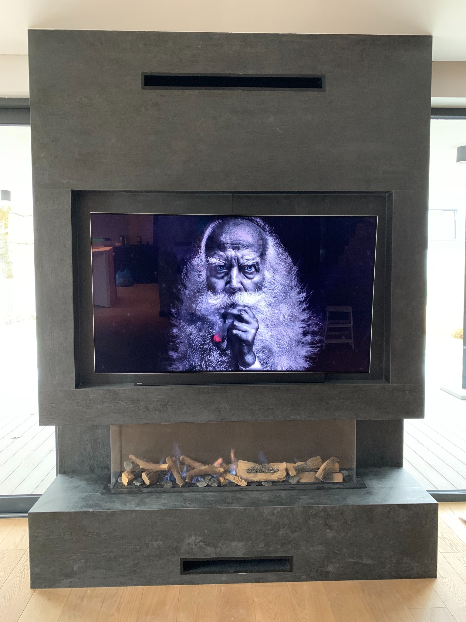Nowoczesny kominek obłożony szarym kamieniem z wbudowanym telewizorem wyświetlającym portret starca z brodą i fajką, poniżej imitacja paleniska z płomieniami.