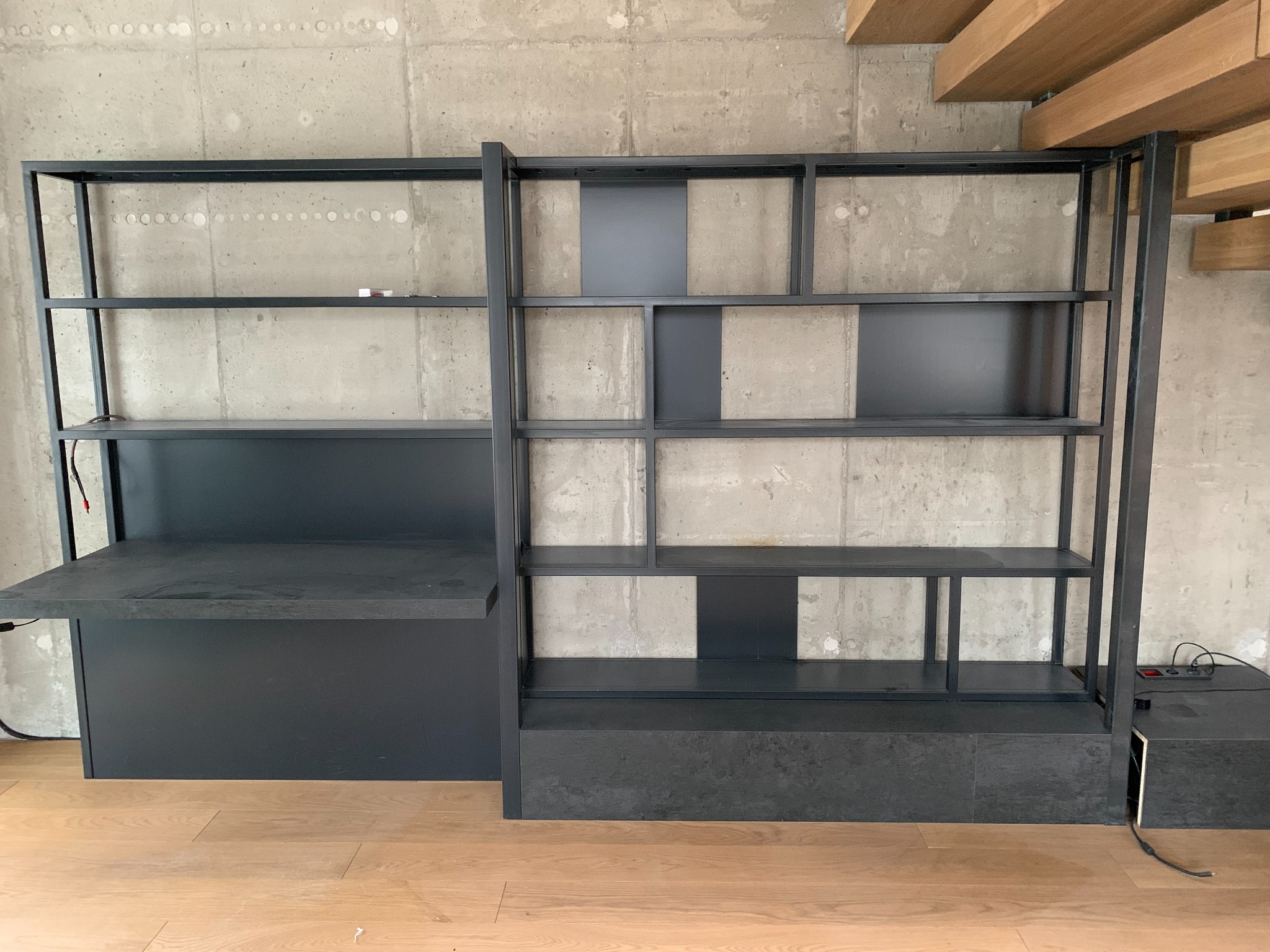 Nowoczesny, minimalistyczny regał z biurkiem, wykonany z ciemnego metalu i płyty, na tle surowej, betonowej ściany z widocznymi śladami po wierceniu i drewnianego sufitu.