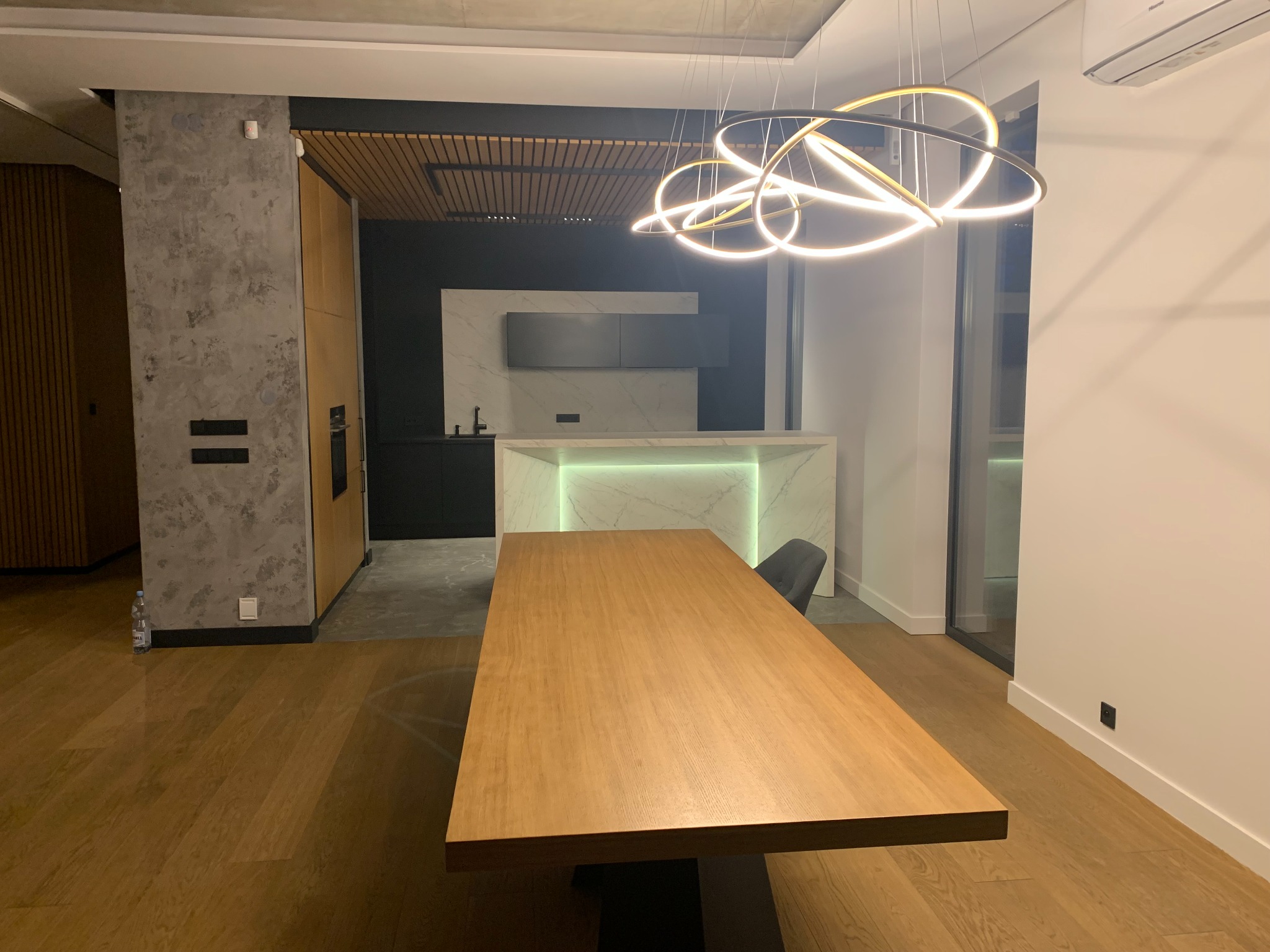 Nowoczesna jadalnia z drewnianym stołem, okrągłą lampą LED i minimalistyczną kuchnią w tle z marmurowym blatem podświetlonym ledami.