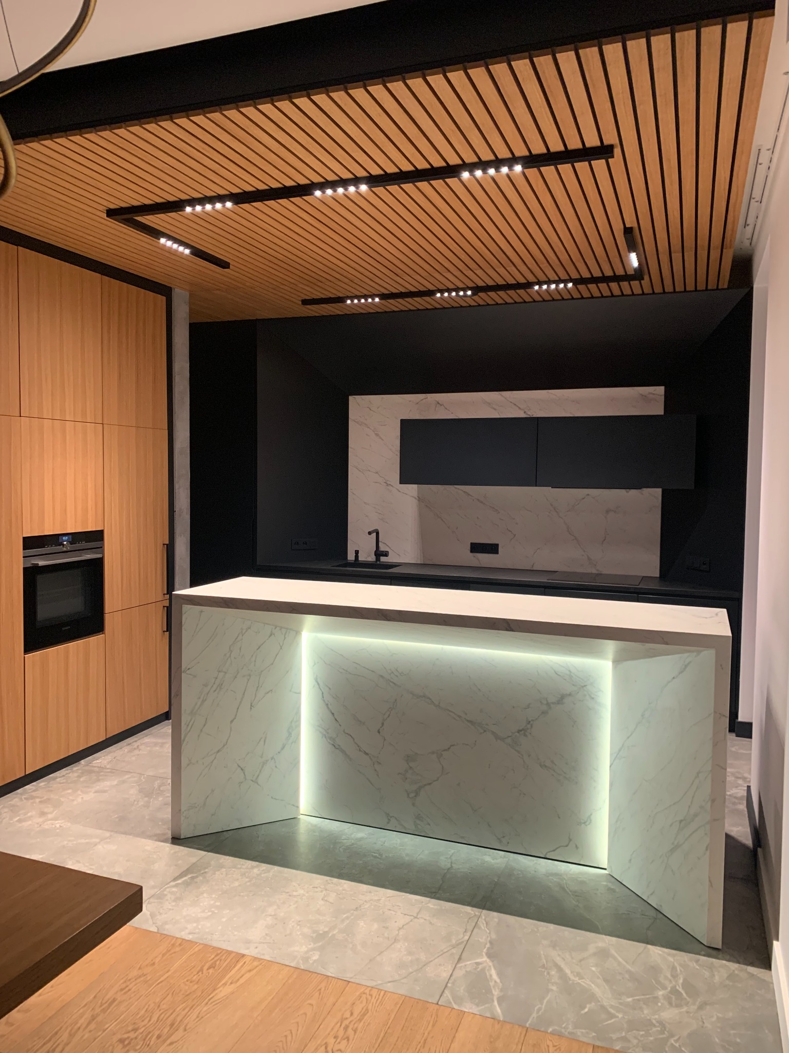 Nowoczesna kuchnia z wyspą oświetloną od spodu, drewnianymi szafkami i sufitem z drewnianych listew z oświetleniem punktowym.