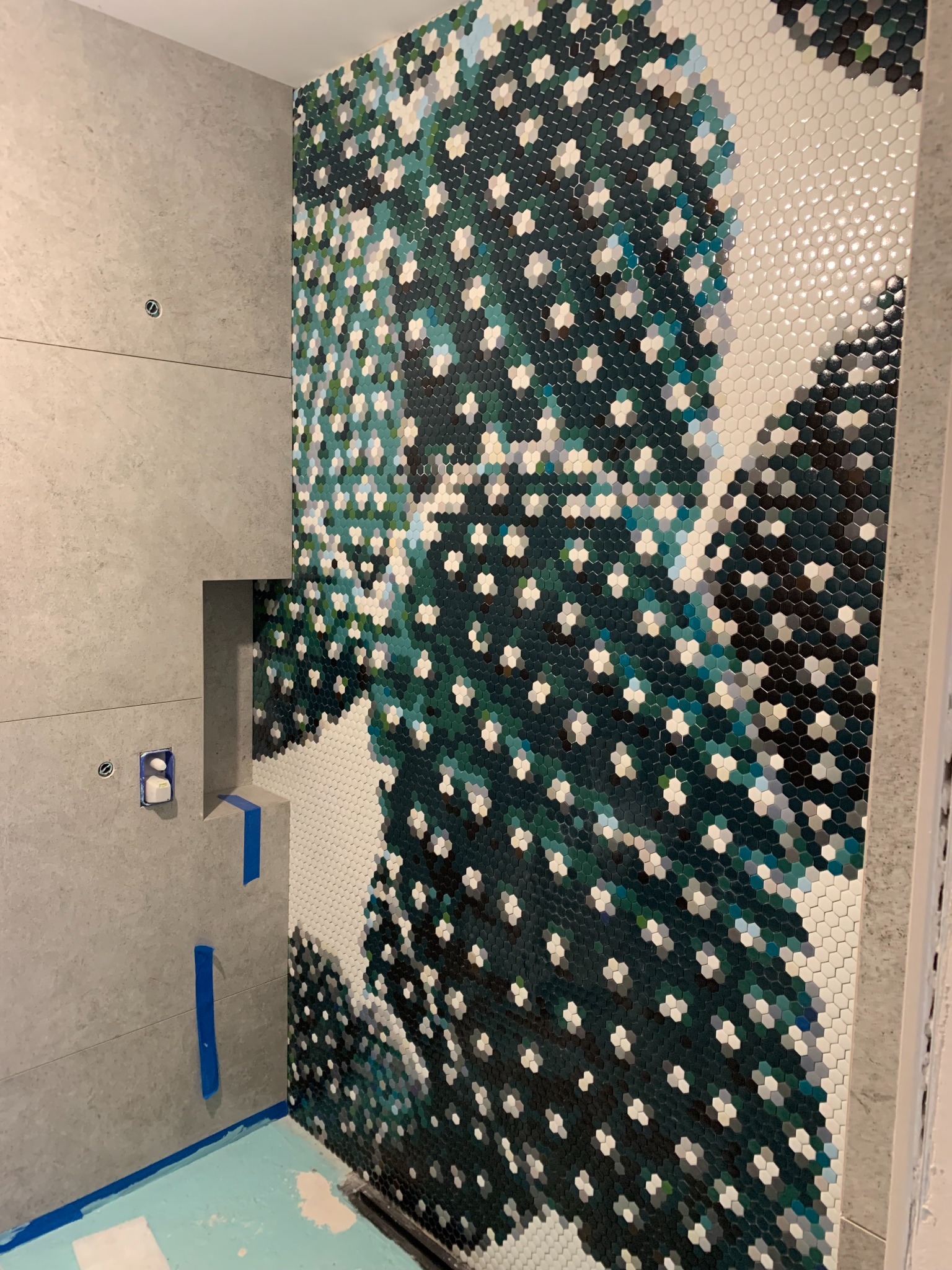 Ściana łazienki w trakcie wykańczania z mozaiką heksagonalną w odcieniach zieleni, bieli i czerni, tworząca wzór przypominający roślinność. Widoczne fragmenty szarej płytki ceramicznej i niebieska...