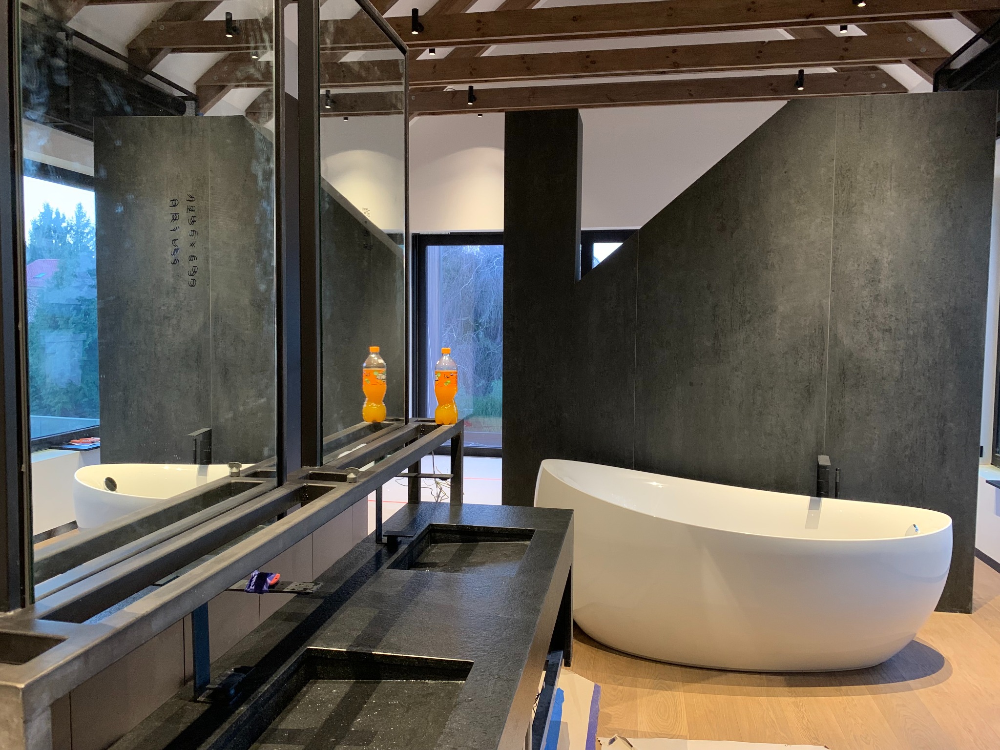 Nowoczesna łazienka z czarnymi, teksturowanymi płytami ściennymi, wolnostojącą białą wanną i podwójną umywalką z czarnego kamienia. Dwie butelki napoju na blacie.