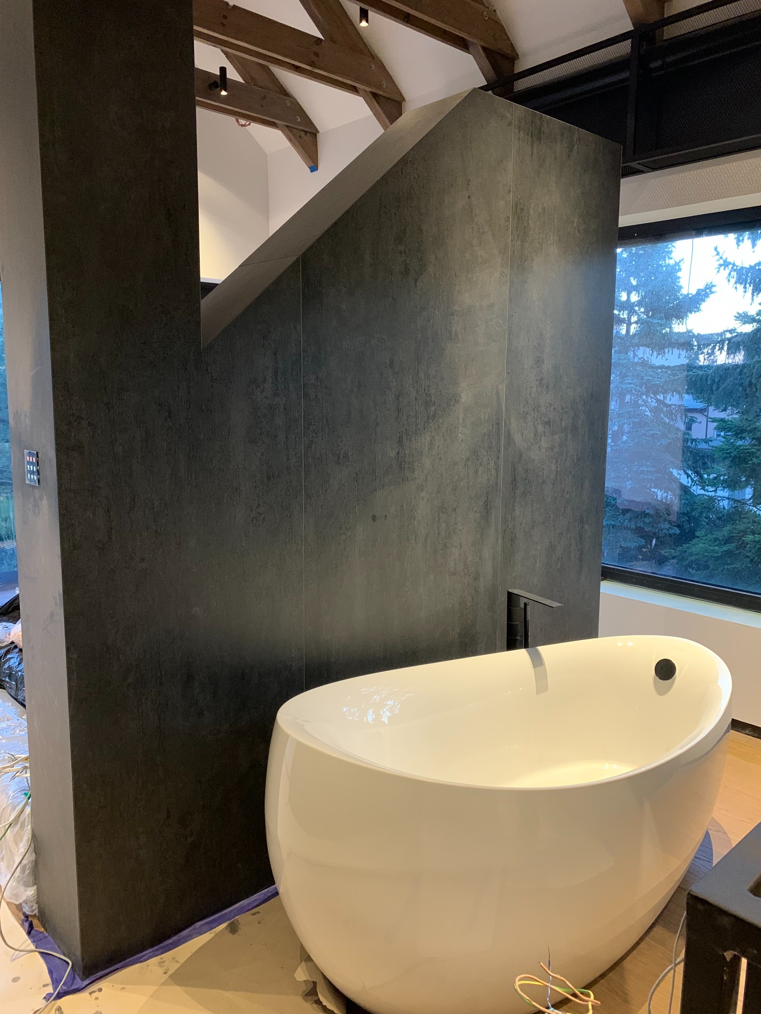 Nowoczesna łazienka w trakcie wykończenia: wolnostojąca wanna, ciemnoszare płytki imitujące beton na ścianach, drewniany strop z oświetleniem punktowym, okno z widokiem na zieleń, zabezpieczenia...