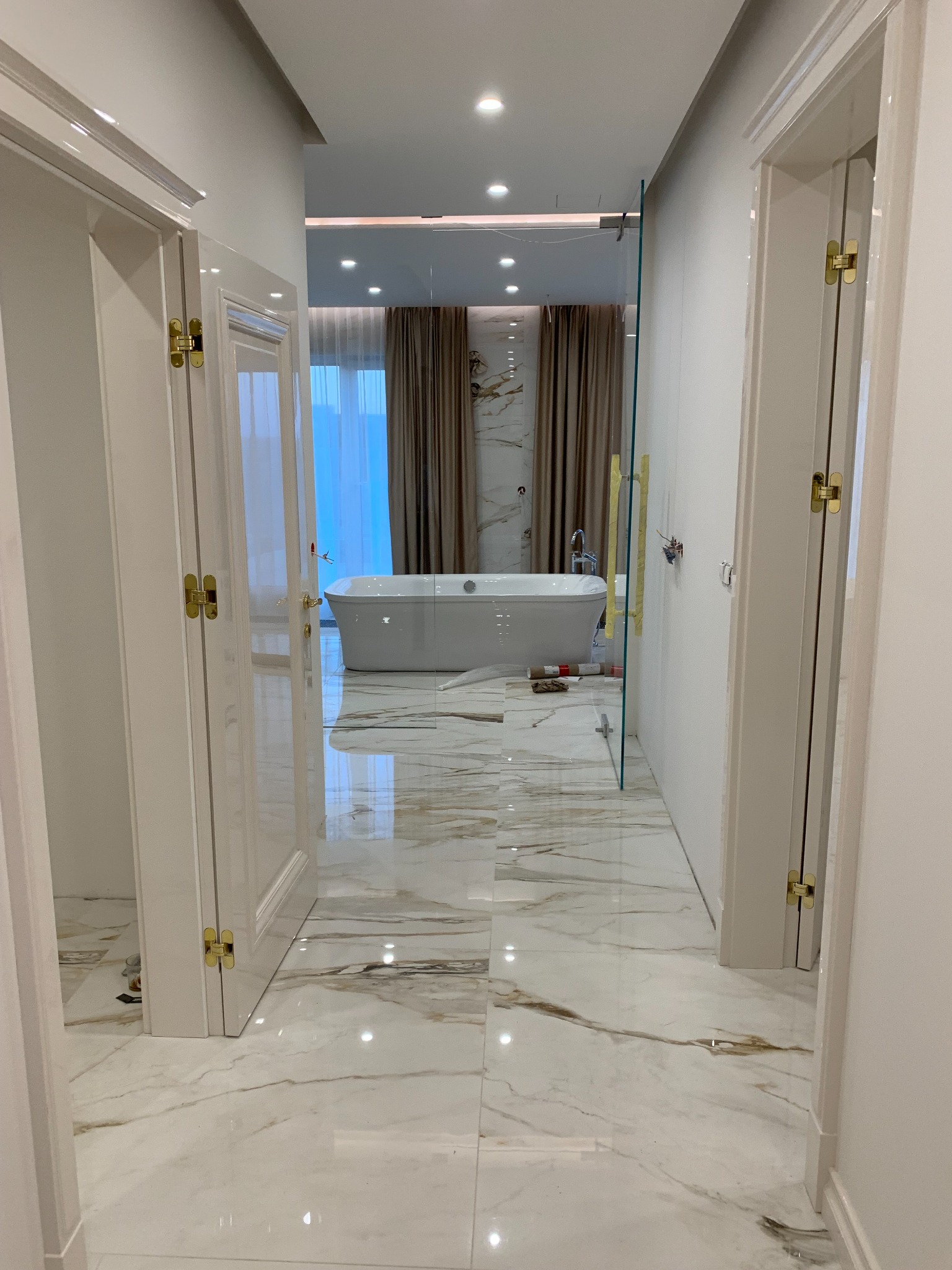Luksusowa łazienka w trakcie remontu, widok z korytarza przez otwarte białe drzwi, marmurowe płytki podłogowe w odcieniach bieli i złota, wolnostojąca wanna, szklana ścianka prysznicowa w trakcie...