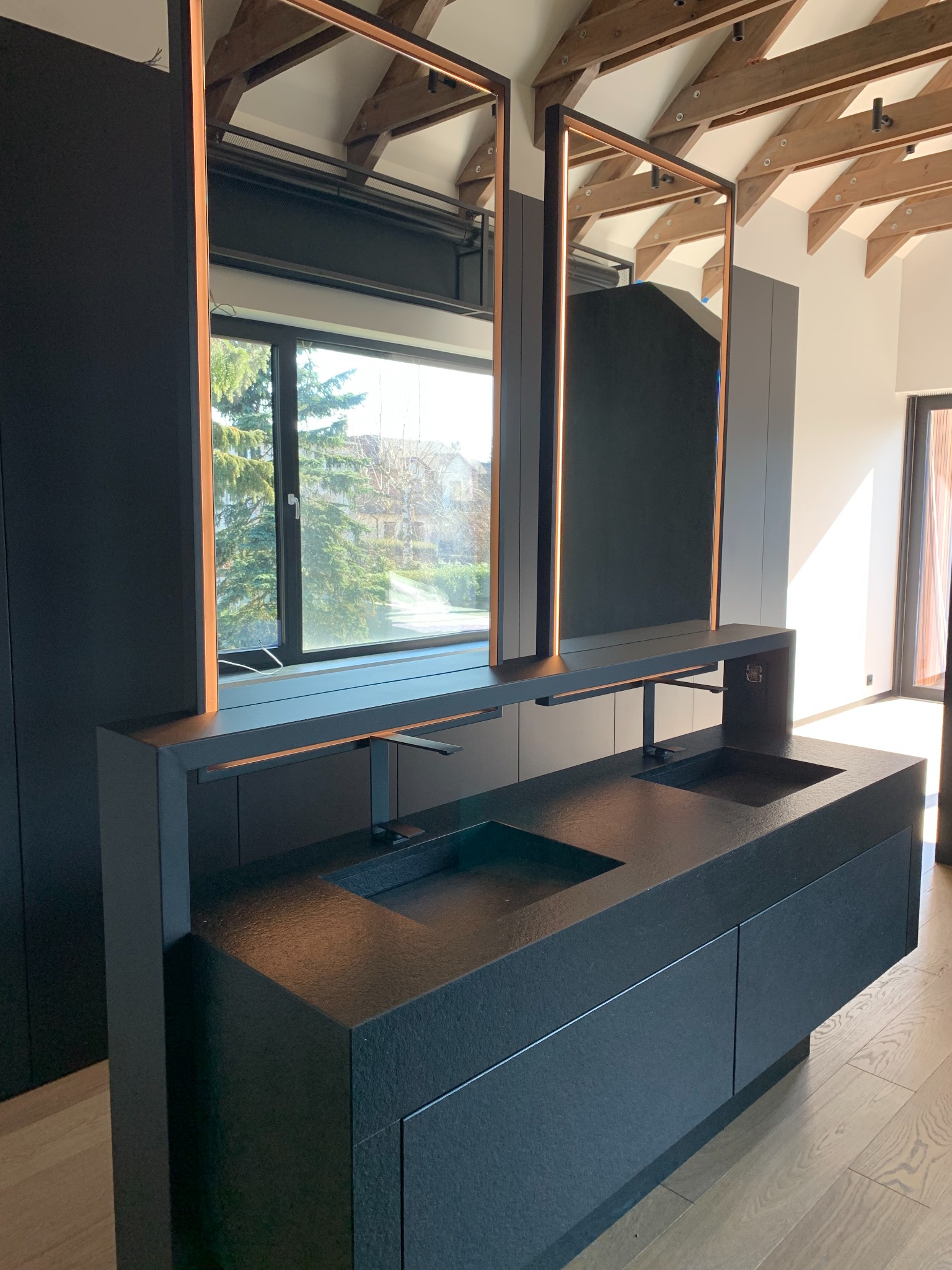 Nowoczesna czarna łazienka z dwiema umywalkami z kamienia, podświetlanymi lustrami i drewnianym sufitem w tle.