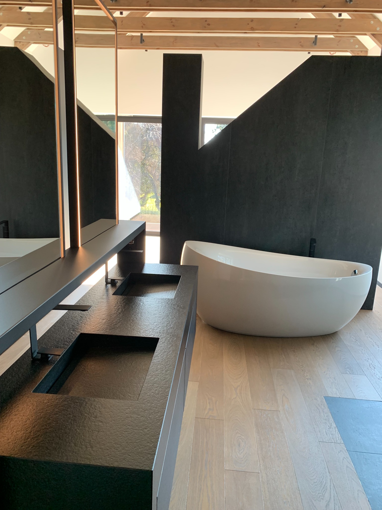 Nowoczesna łazienka z podwójną, czarną umywalką z kamienia, wolnostojącą białą wanną i drewnianą podłogą w jasnym odcieniu, oświetlona naturalnym światłem.