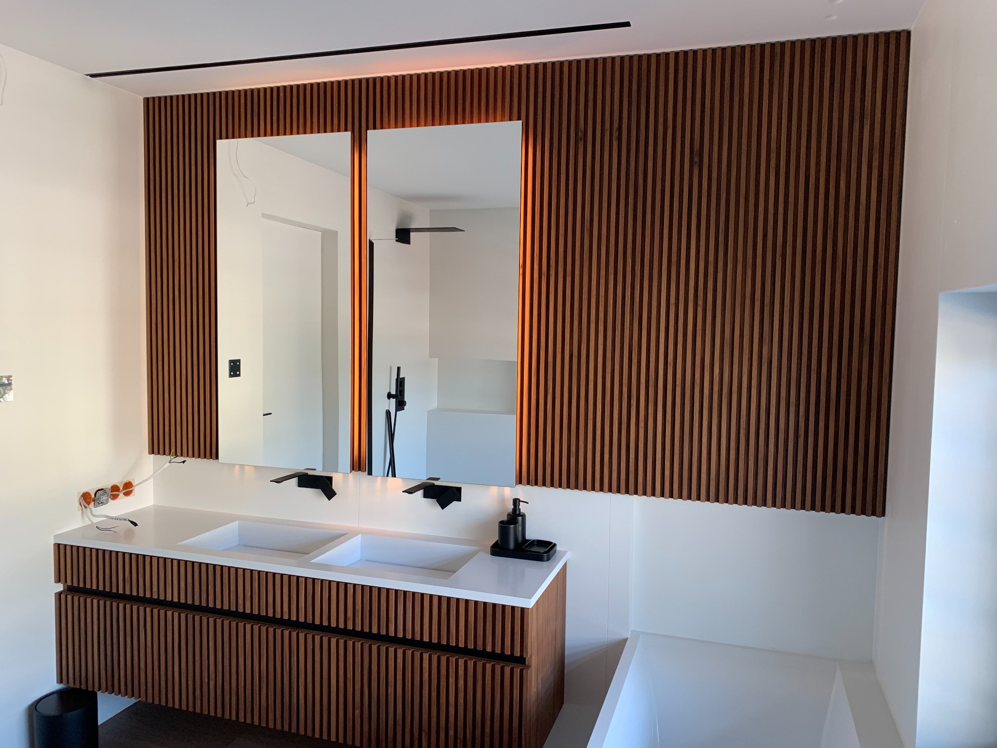 Nowoczesna łazienka z drewnianą ścianą z pionowych listew, podświetlanymi lustrami, białą umywalką z dwiema misami i czarną armaturą.