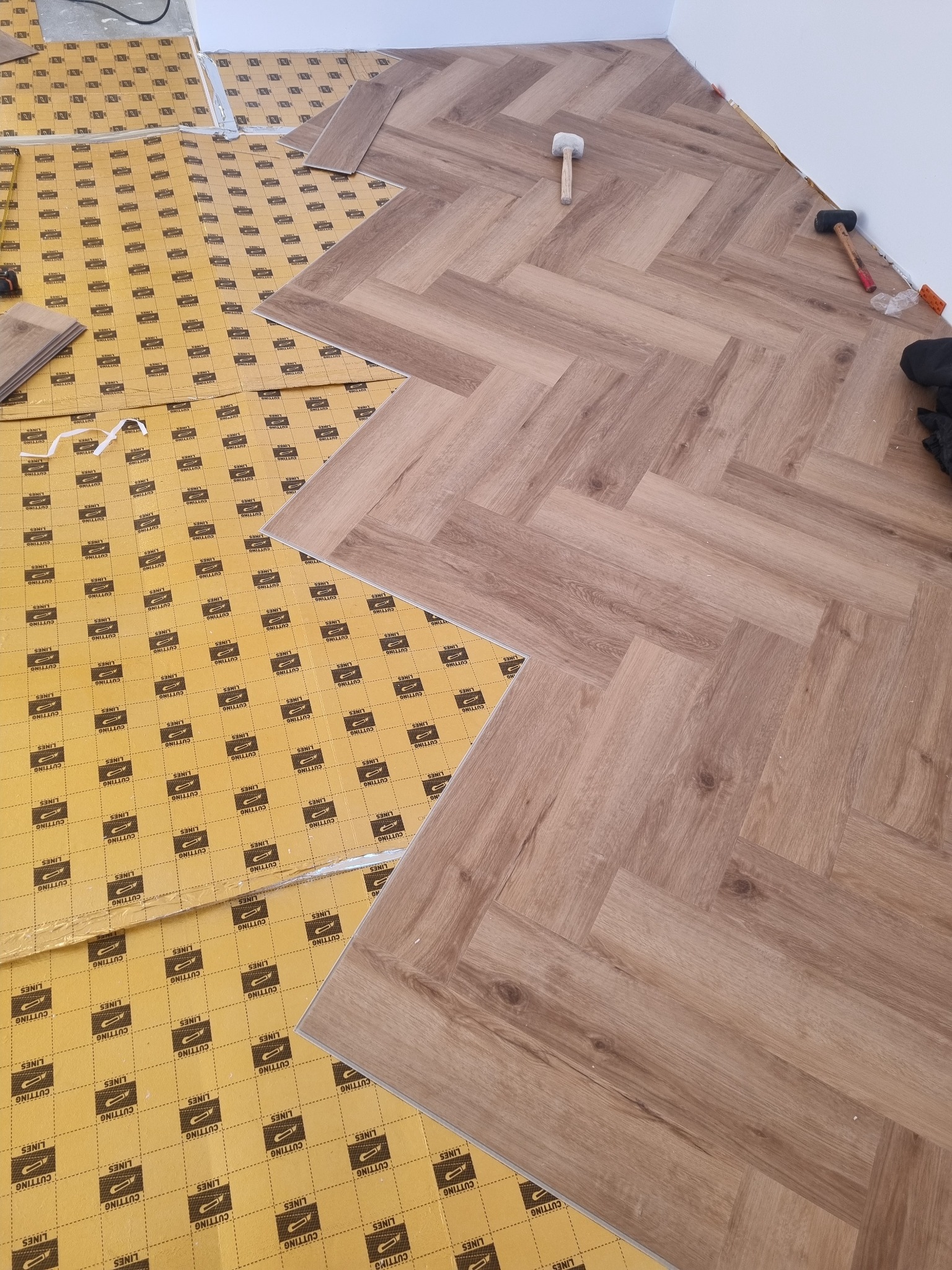 Układanie podłogi z paneli w jodełkę, widoczne narzędzia: gumowy młotek i kliny dystansowe, podkład pod panele z nadrukiem, fragment ściany w rogu pomieszczenia.