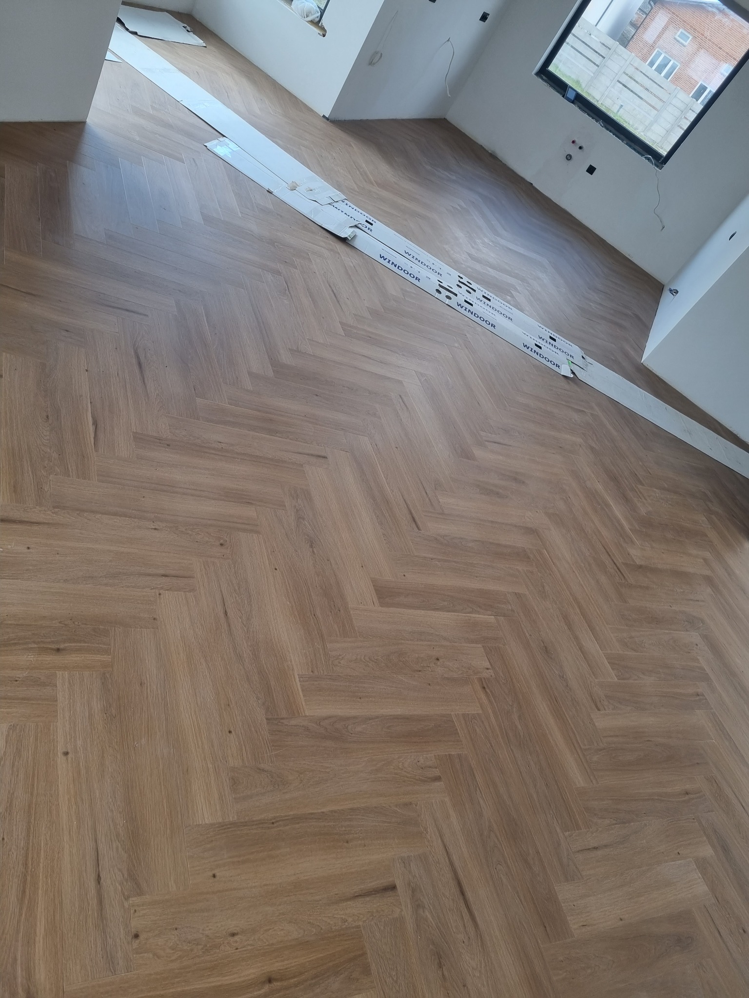 Ukończona podłoga z parkietu ułożonego we wzór jodełki francuskiej, z widocznymi paskami kartonu ochronnego na środku pomieszczenia, białe ściany i okno w tle.