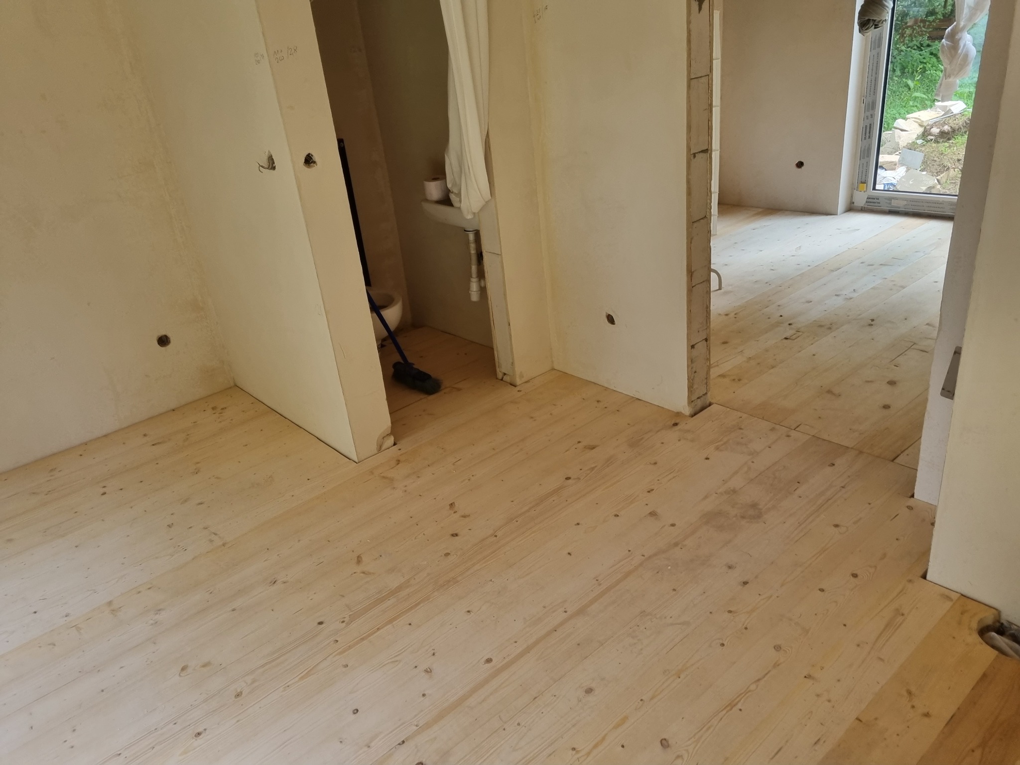 Jasny, surowy parkiet z widocznymi sękami w trakcie układania, widoczne przygotowane ściany i otwarte przejścia do innych pomieszczeń, w tle fragment łazienki z umywalką.
