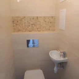 Mała toaleta dla gości z beżowymi płytkami imitującymi kamień, mozaiką nad zabudową stelaża WC, umywalką narożną i chromowaną armaturą.