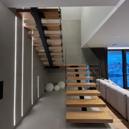 F.H.P Gawor Piotr Gawor - Nowoczesne schody drewniane z czarnymi wspornikami i szklaną balustradą, oświetlone pionowymi liniami LED na ścianie. Minimalistyczny design.