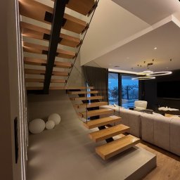 F.H.P Gawor Piotr Gawor - Nowoczesne schody drewniane z czarną konstrukcją i szklanymi panelami, w jasnym wnętrzu z widokiem na zewnątrz. Minimalistyczny design, ciepłe oświetlenie.