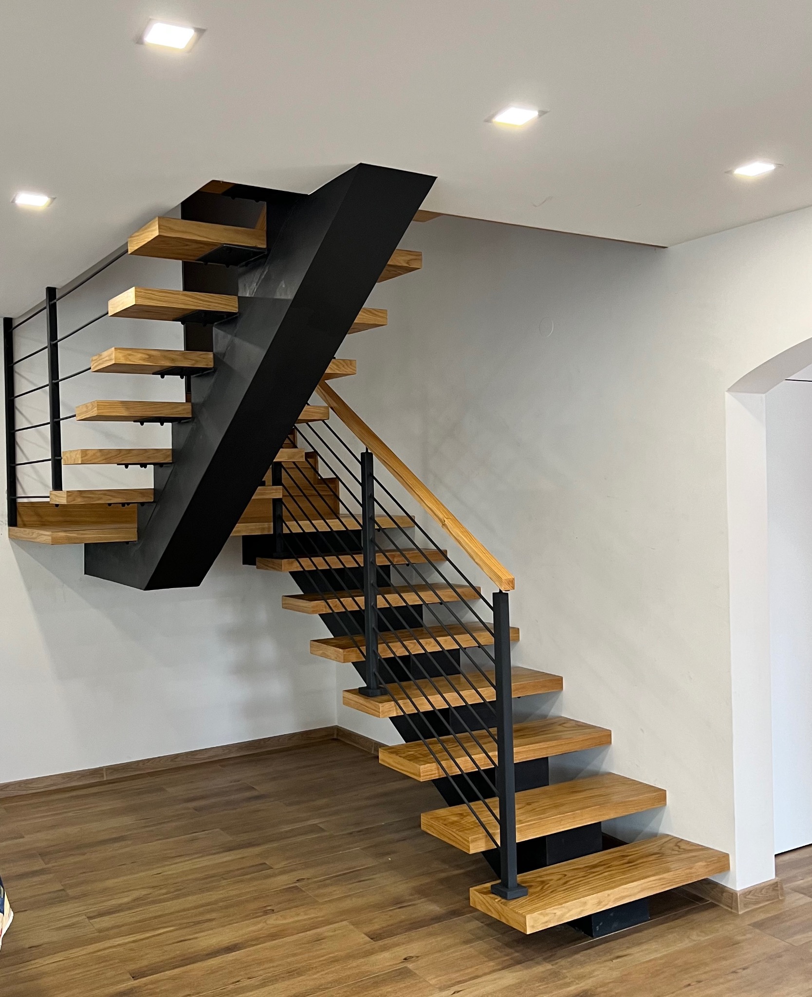 Nowoczesne schody dywanowe z drewnianymi stopniami i czarną metalową konstrukcją, minimalistyczne balustrady z cienkich prętów, jasne wnętrze.