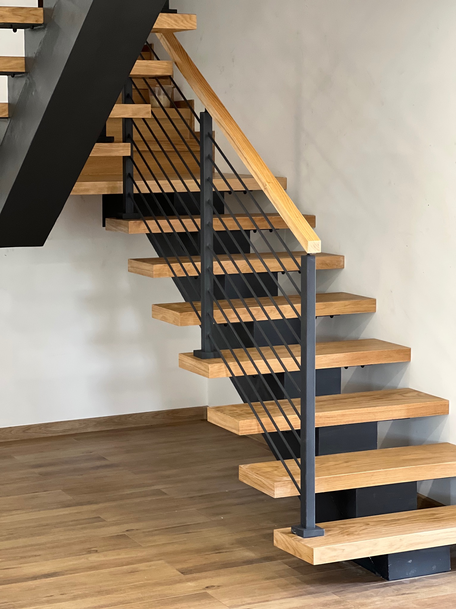 Nowoczesne schody z drewnianymi stopniami, czarnymi wspornikami i metalową balustradą z drewnianą poręczą, na tle jasnej ściany i podłogi z paneli.
