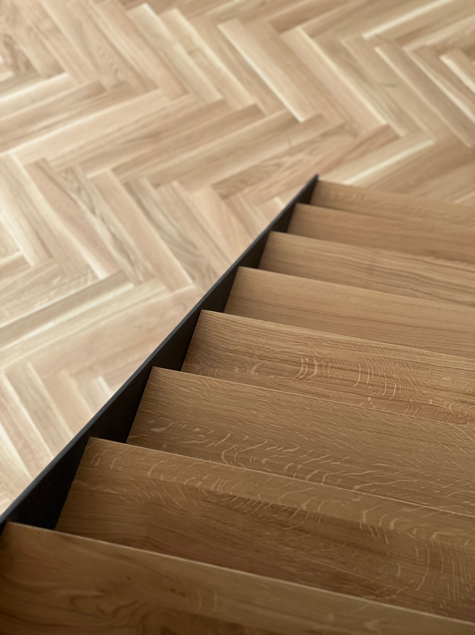 Drewniane schody z czarnym wykończeniem, widok z góry. Parkiet ułożony w jodełkę. Naturalne oświetlenie podkreśla teksturę drewna.