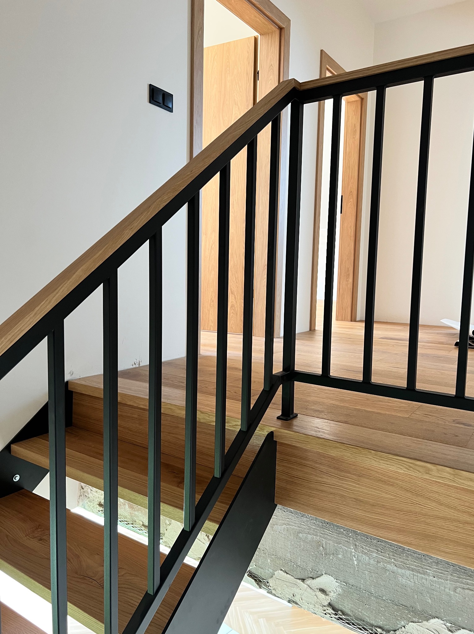 Nowoczesne schody z drewnianymi stopniami i czarną, metalową balustradą. Widok na otwarte drzwi w tle. Detal wykończenia schodów w surowym stanie.