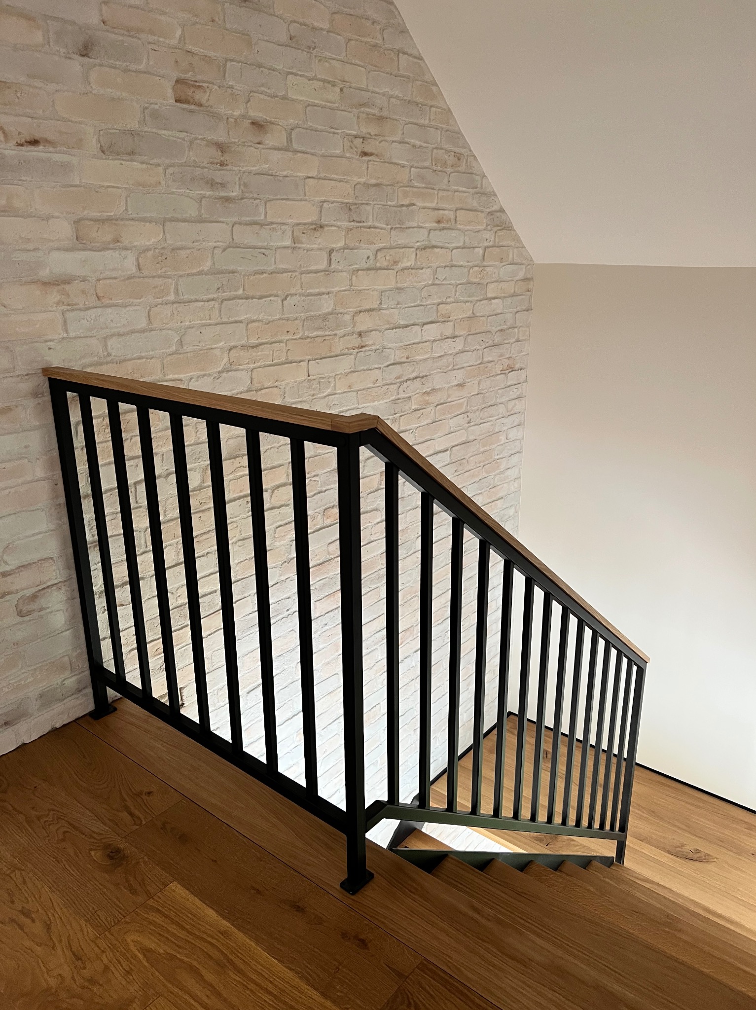 Nowoczesne schody z drewnianymi stopniami i metalową, czarną balustradą z drewnianym poręczem, na tle ściany z cegły w odcieniach bieli i beżu.