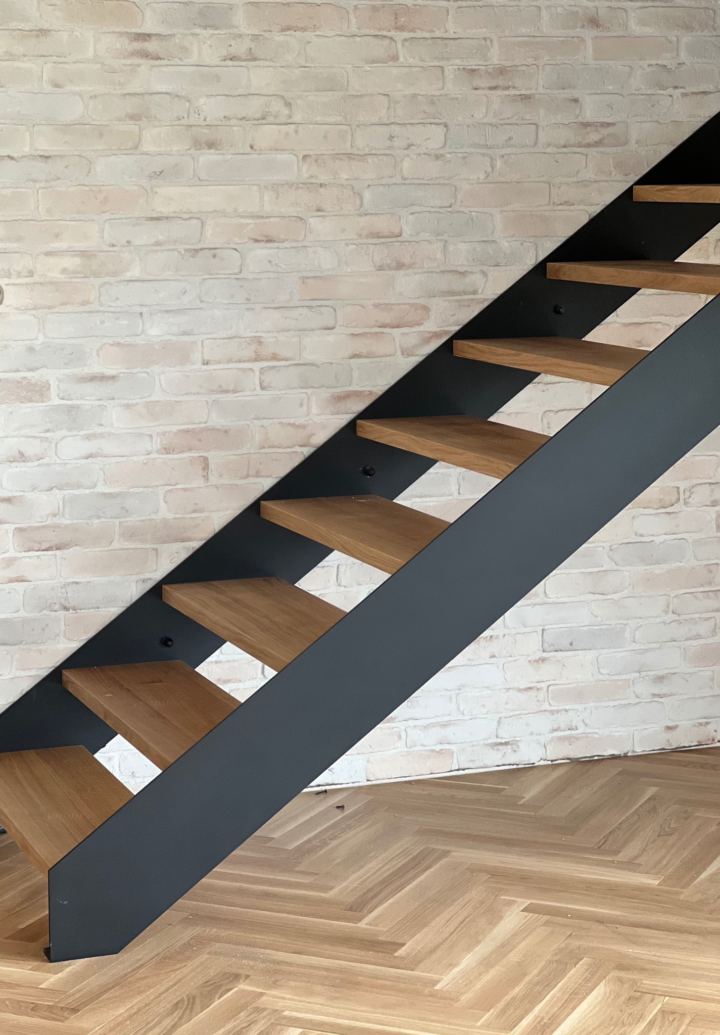 Nowoczesne schody z drewnianymi stopniami i metalową konstrukcją na tle ceglanej ściany i parkietu w jodełkę. Minimalistyczny design.
