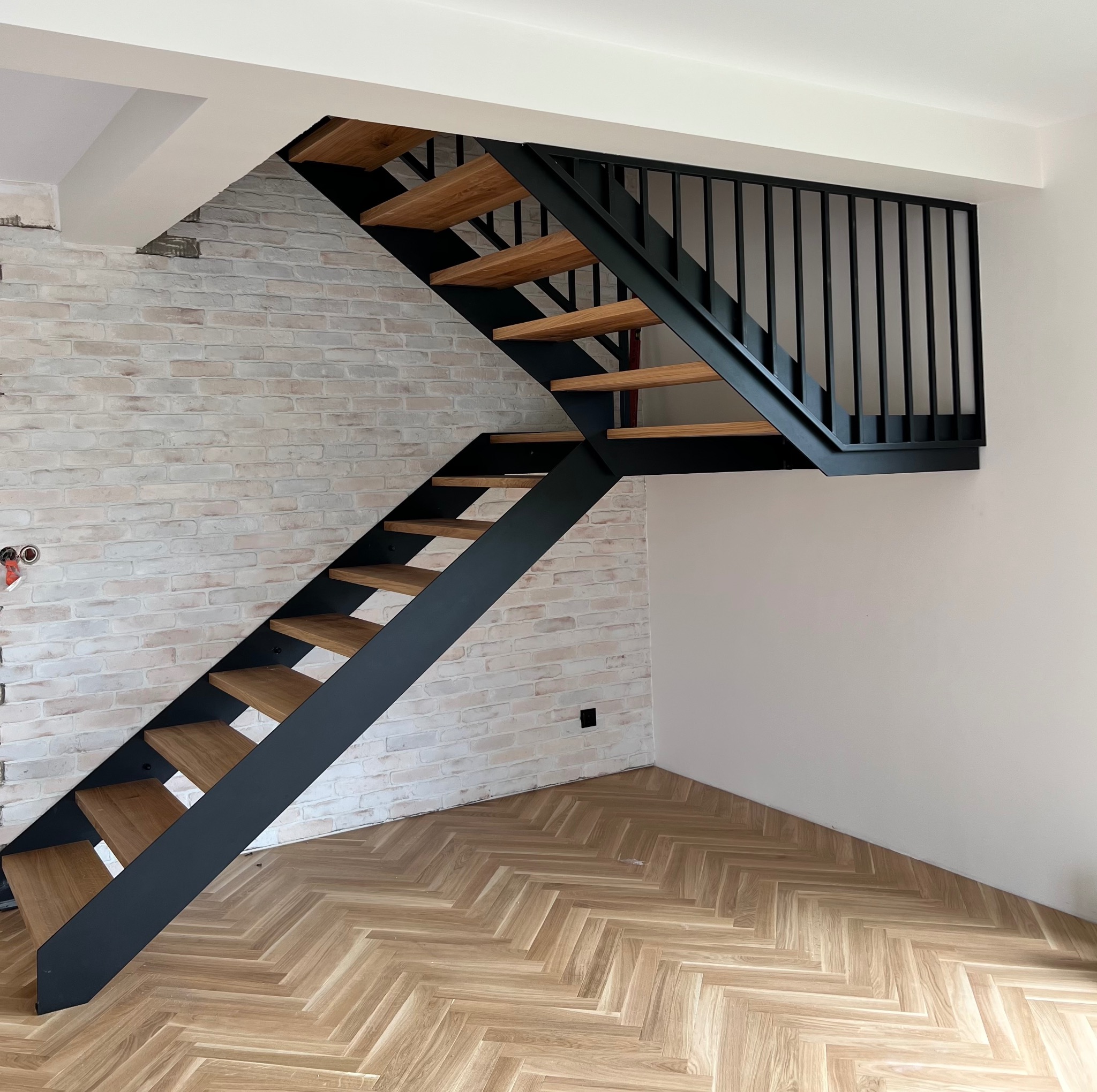Nowoczesne schody metalowe z drewnianymi stopniami, czarna konstrukcja, jasna cegła na ścianie, parkiet w jodełkę. Minimalistyczny design i solidne wykonanie.