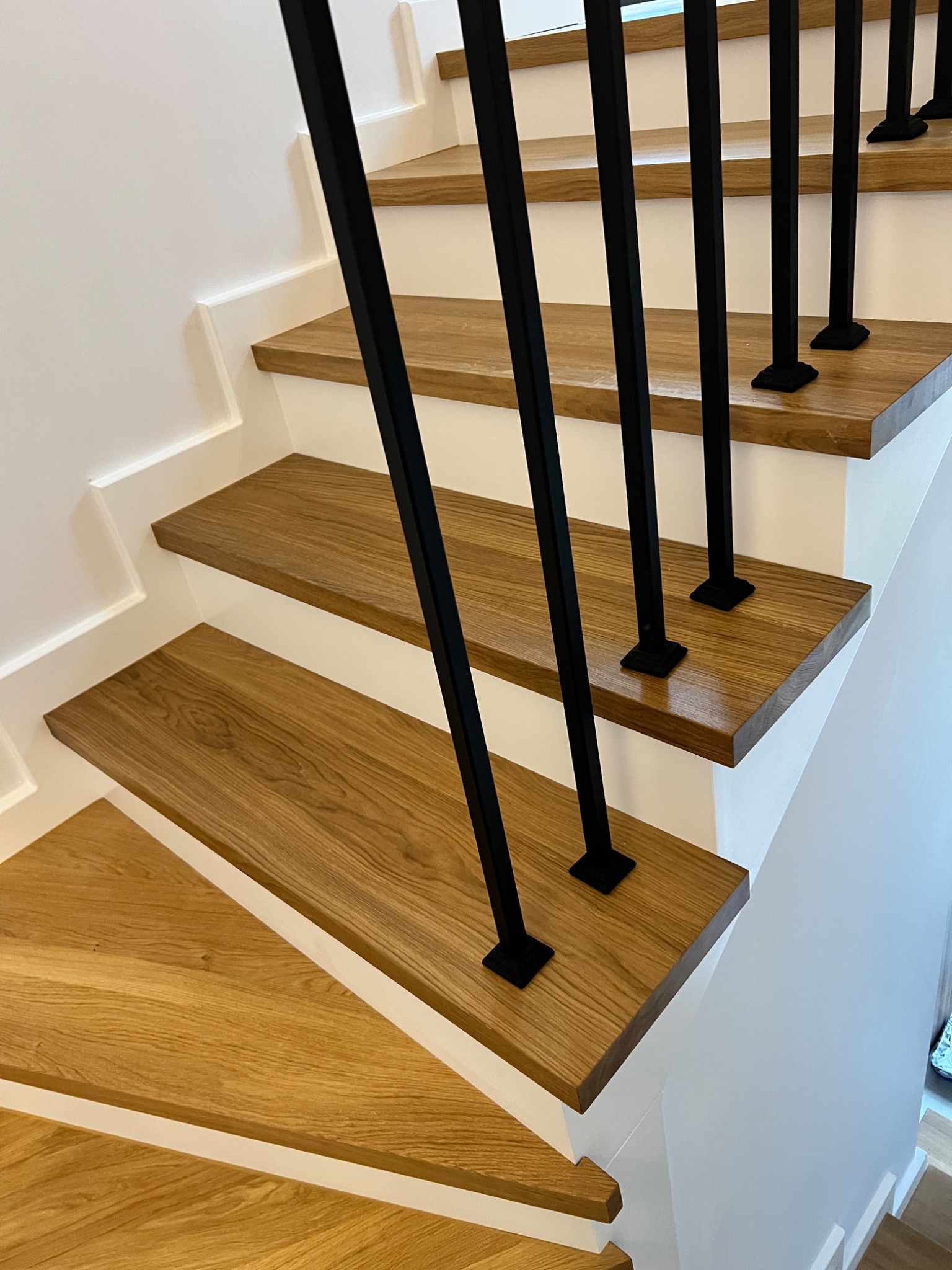 Jasne, dębowe schody z czarnymi, metalowymi słupkami balustrady. Minimalistyczny design, białe ściany. Widok z góry na fragment schodów.