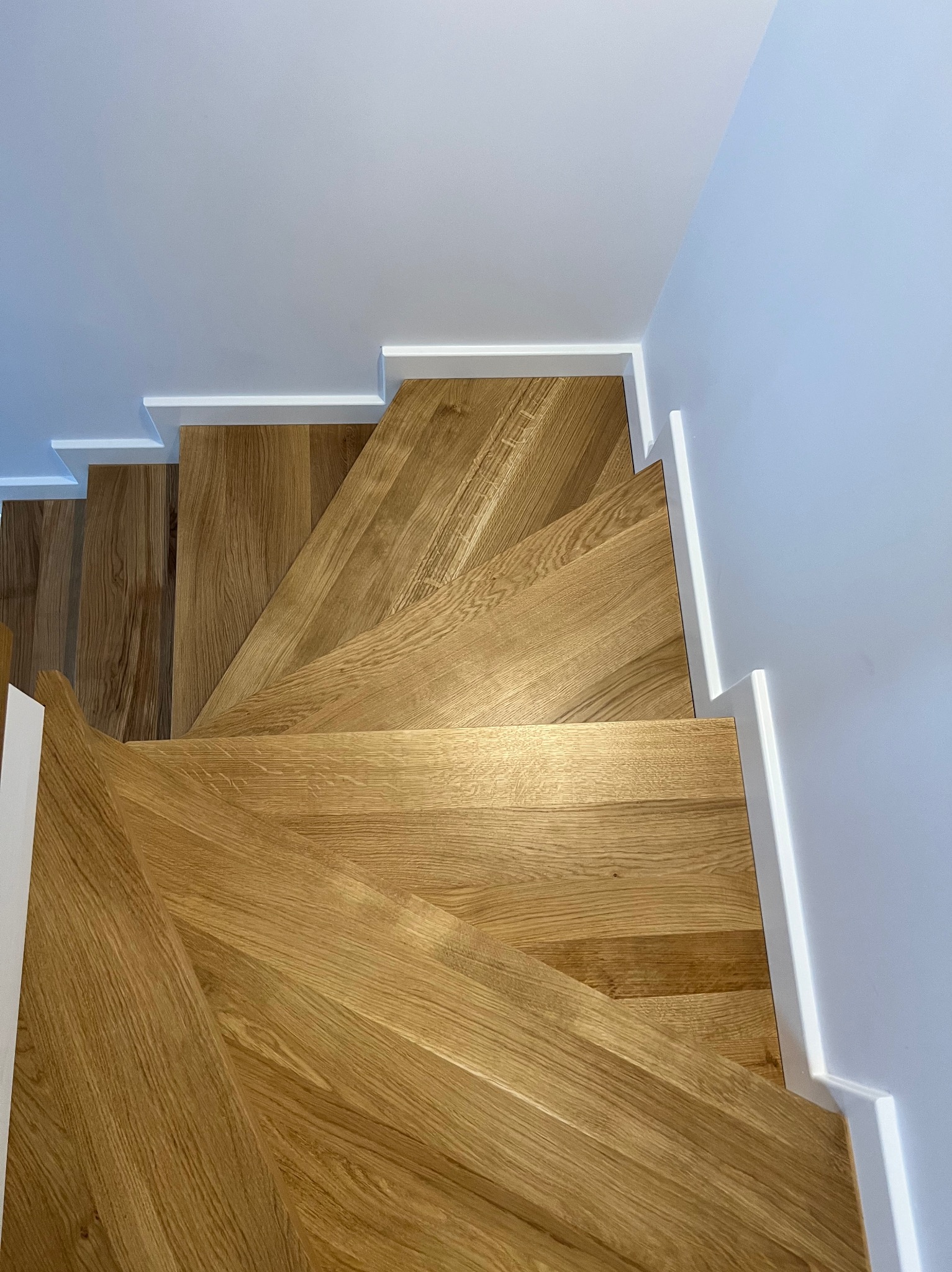 Nowoczesne schody drewniane z jasnego drewna, widok z góry. Minimalistyczne białe wykończenie ścian i listew przypodłogowych. Naturalne oświetlenie podkreśla strukturę drewna.