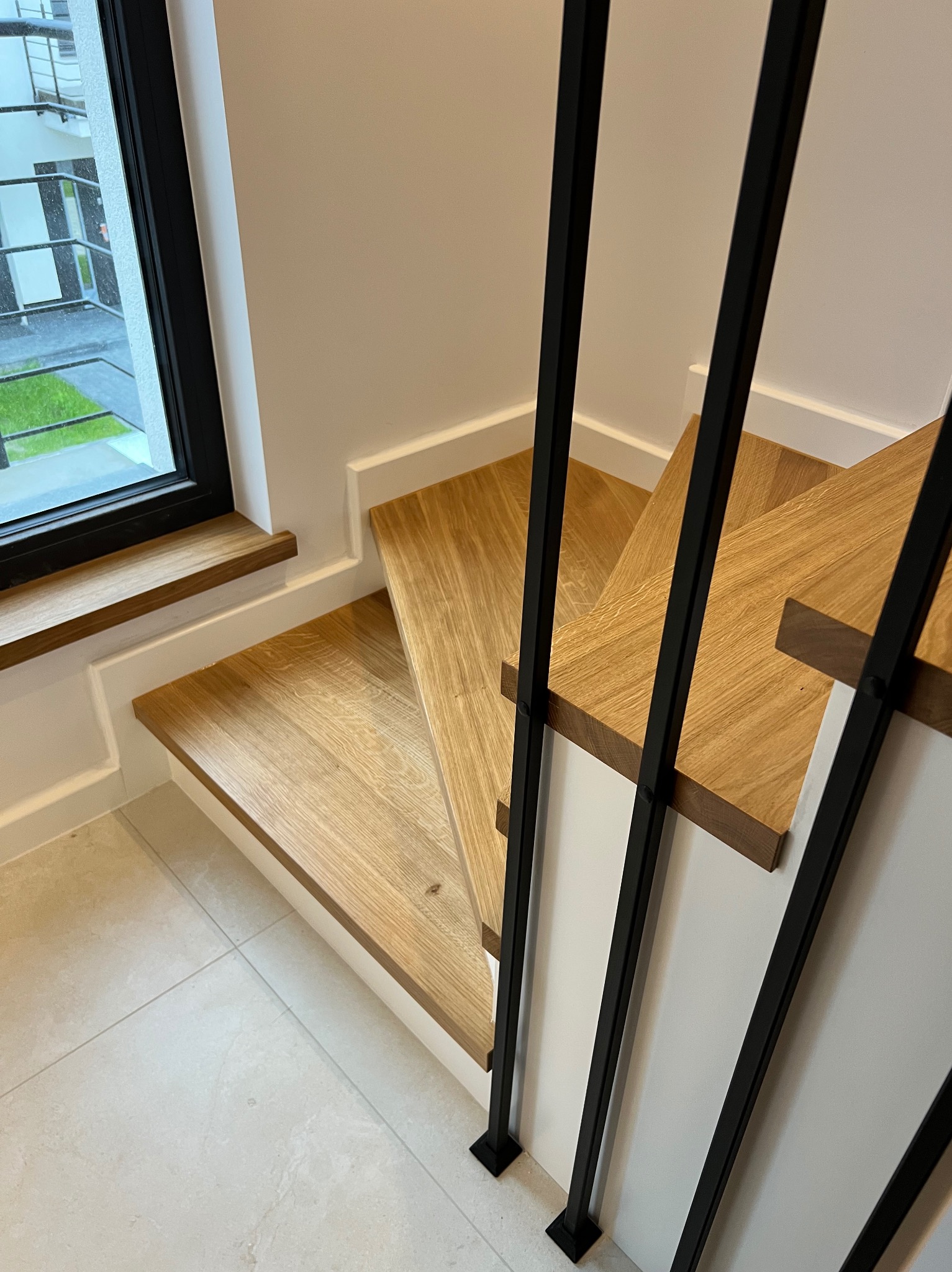 Nowoczesne schody z drewnianymi stopniami i czarnymi, metalowymi poręczami, widok z góry, z oknem w tle. Minimalistyczny design w jasnym wnętrzu.
